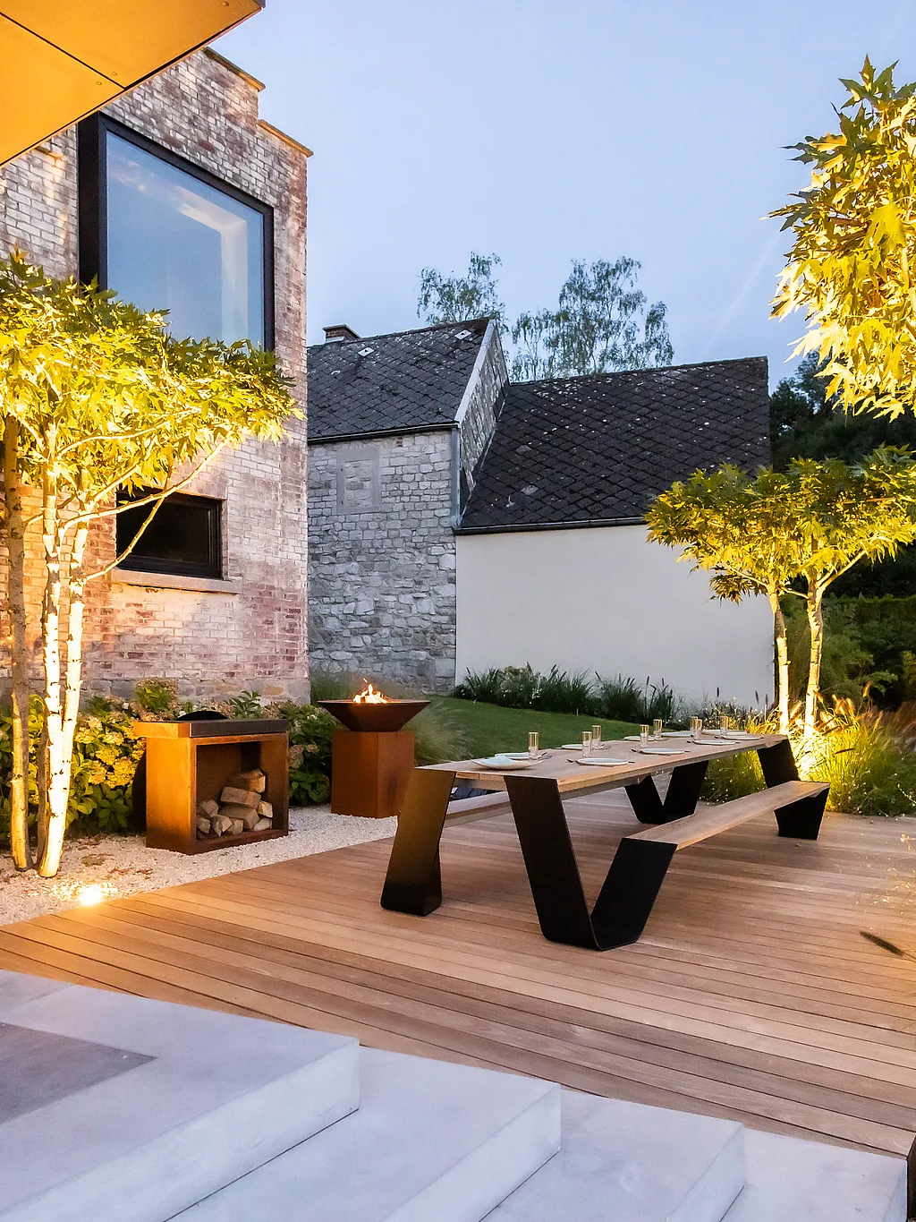 Vue en soirée sur un jardin en terrasse à côté d’une maison en brique et pierre, avec des arbres sculptés éclairés chaleureusement, une longue table à manger en bois avec des bancs sur une terrasse, et un coin salon brillant au loin.