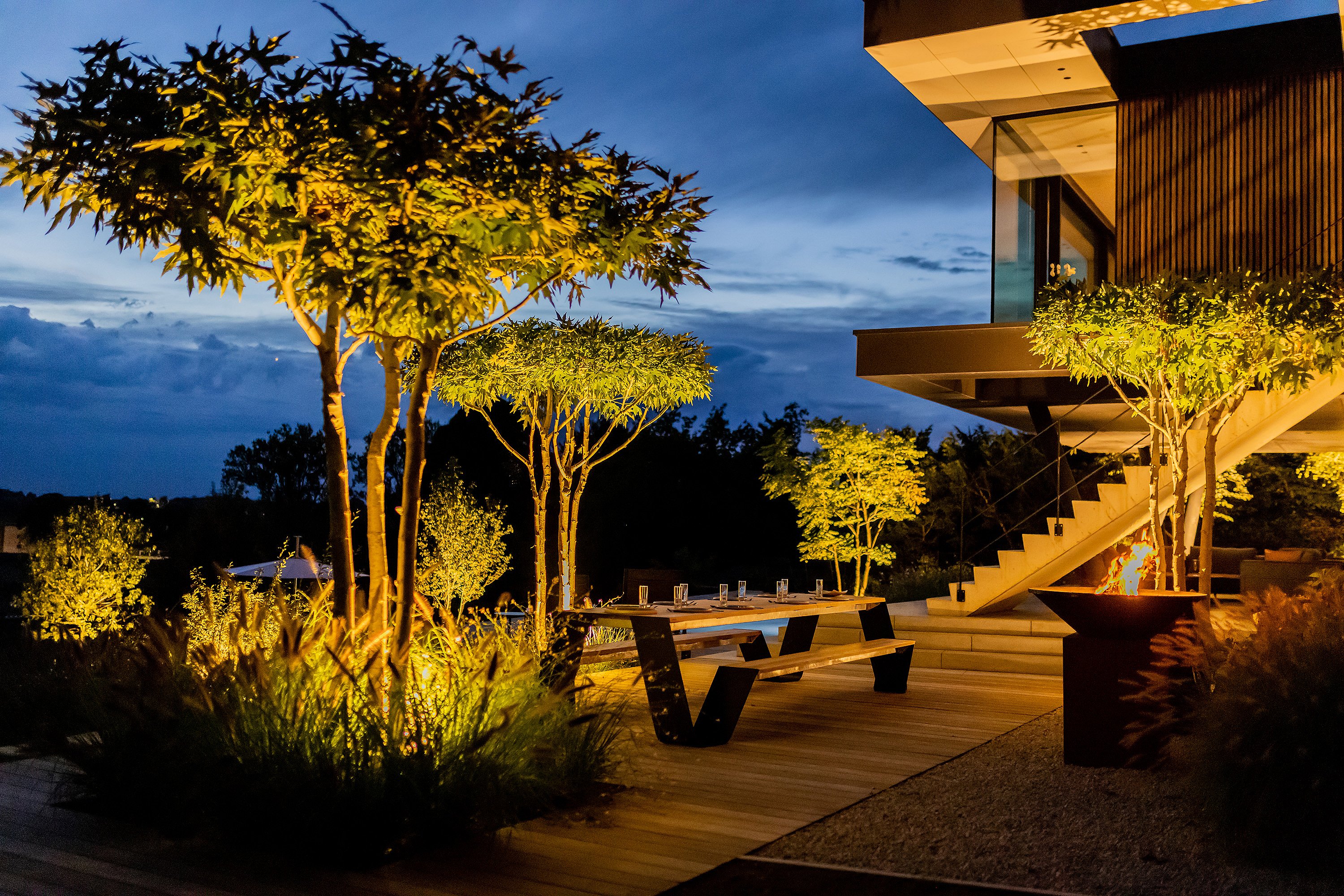 Nachtblick auf die Speiseterrasse mit beleuchteten Bäumen, einem langen Holztisch zum Abendessen, einer brennenden Feuerschale und dem modernen Haus, das neben dem Garten leuchtet.