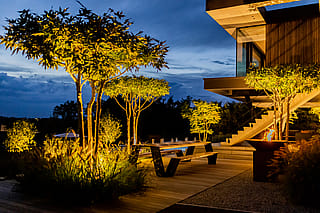 Nachtblick auf die Speiseterrasse mit beleuchteten Bäumen, einem langen Holztisch zum Abendessen, einer brennenden Feuerschale und dem modernen Haus, das neben dem Garten leuchtet.