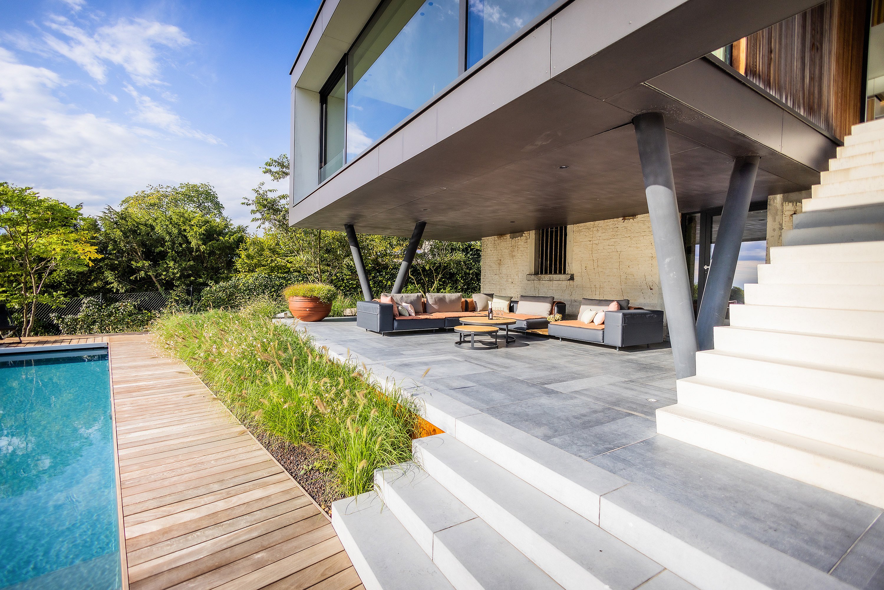 Überdachte Außenlounge unter einem freitragenden Obergeschoss, ausgestattet mit Sofas aus grauem Walross und runden Couchtischen neben einem Pool und einer breiten weißen Treppe.