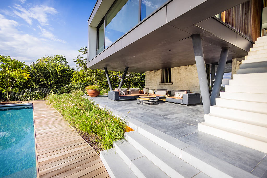 Überdachte Außenlounge unter einem freitragenden Obergeschoss, ausgestattet mit Sofas aus grauem Walross und runden Couchtischen neben einem Pool und einer breiten weißen Treppe.