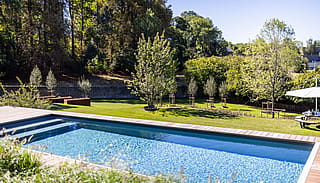 Langer rechteckiger Swimmingpool mit Holzterrasse, abfallender Rasenfläche, jungen Bäumen und einem runden Picknicktisch unter einem weißen Sonnenschirm am Rand des Gartens.