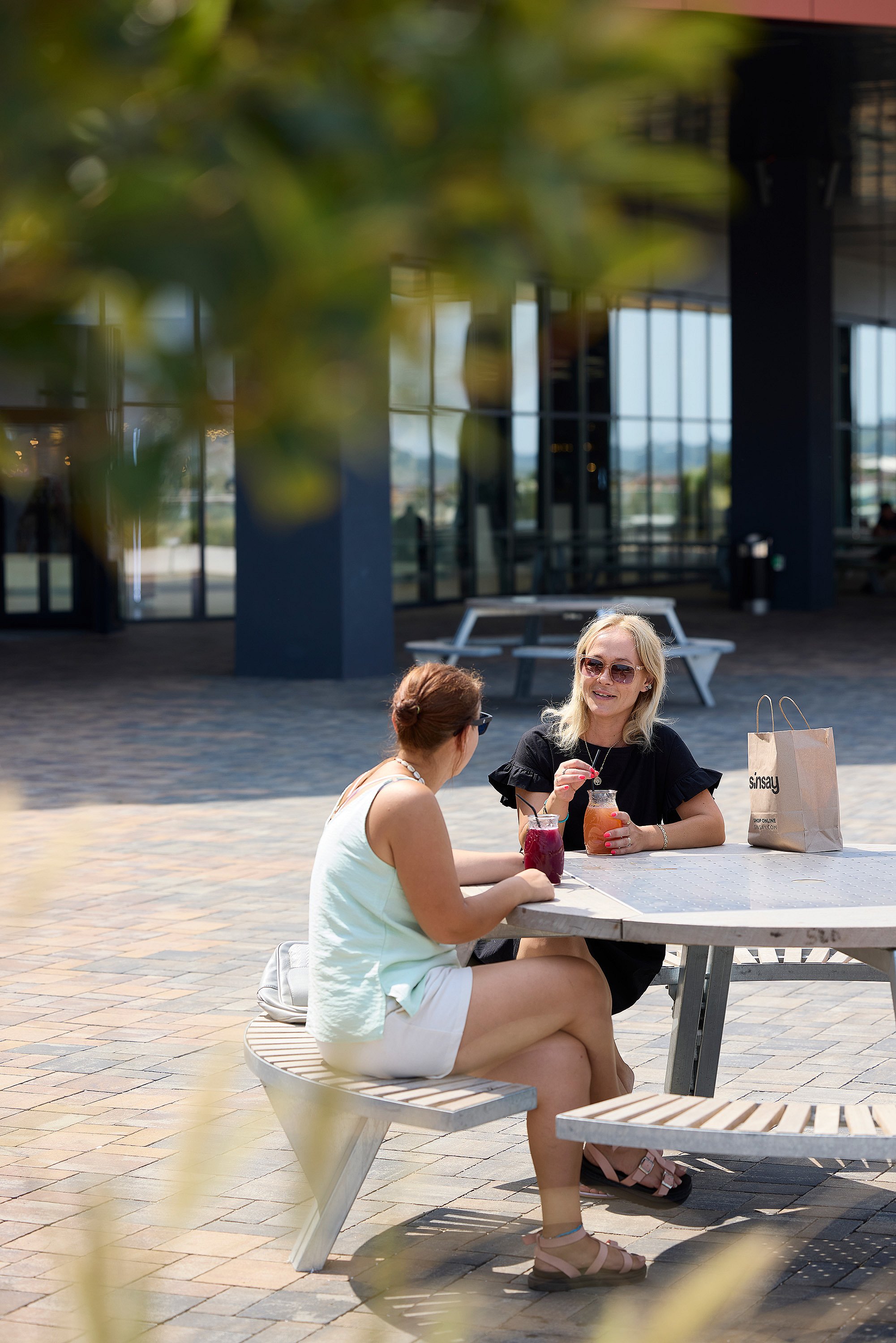 Two women sitting at a light-colored round gargantua picnic table with slatted benches, drinking colorful beverages outside the mall.