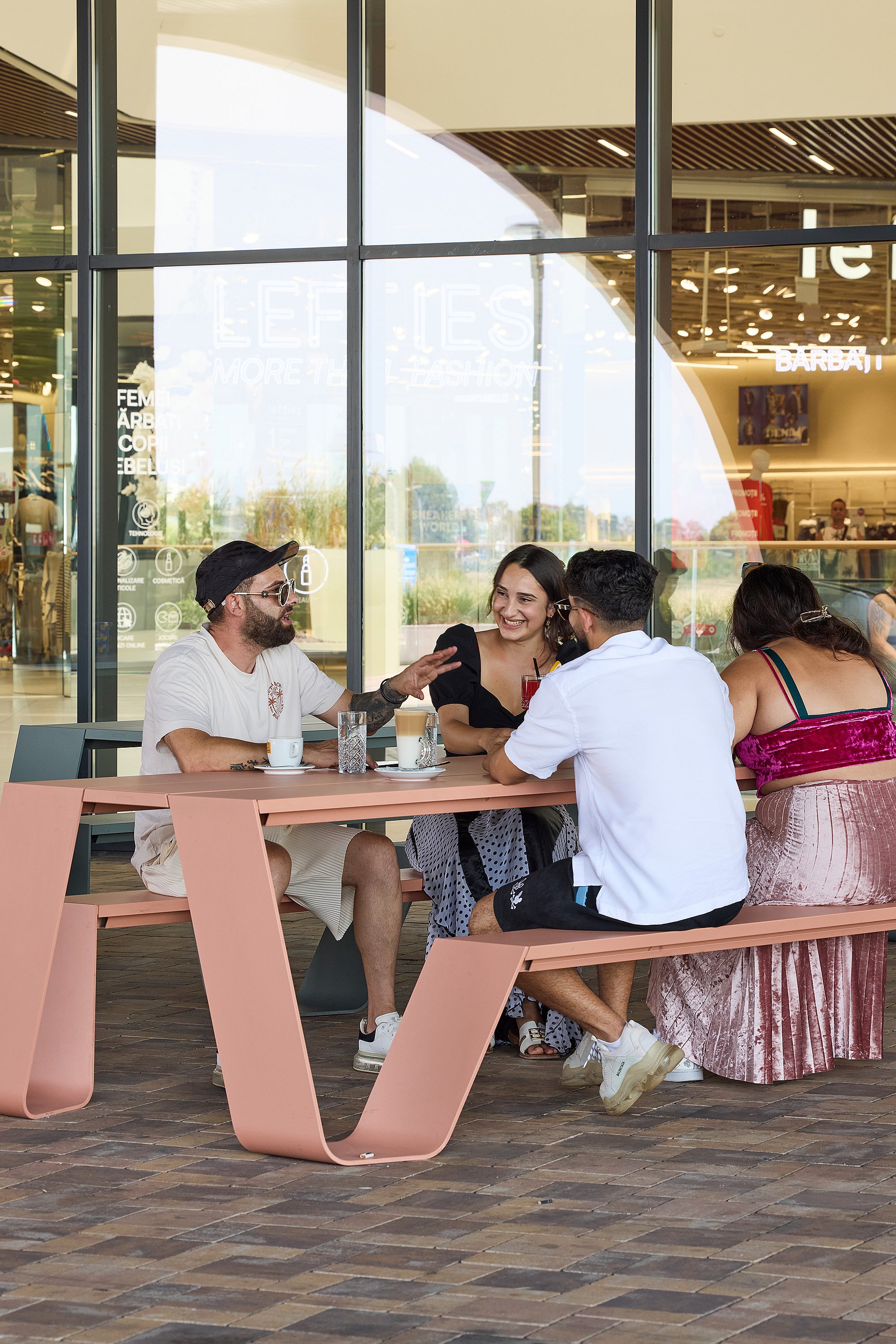 Four friends chatting around a salmon-pink hopper picnic table in front of tall mall windows, with coffee and drinks on the tabletop
