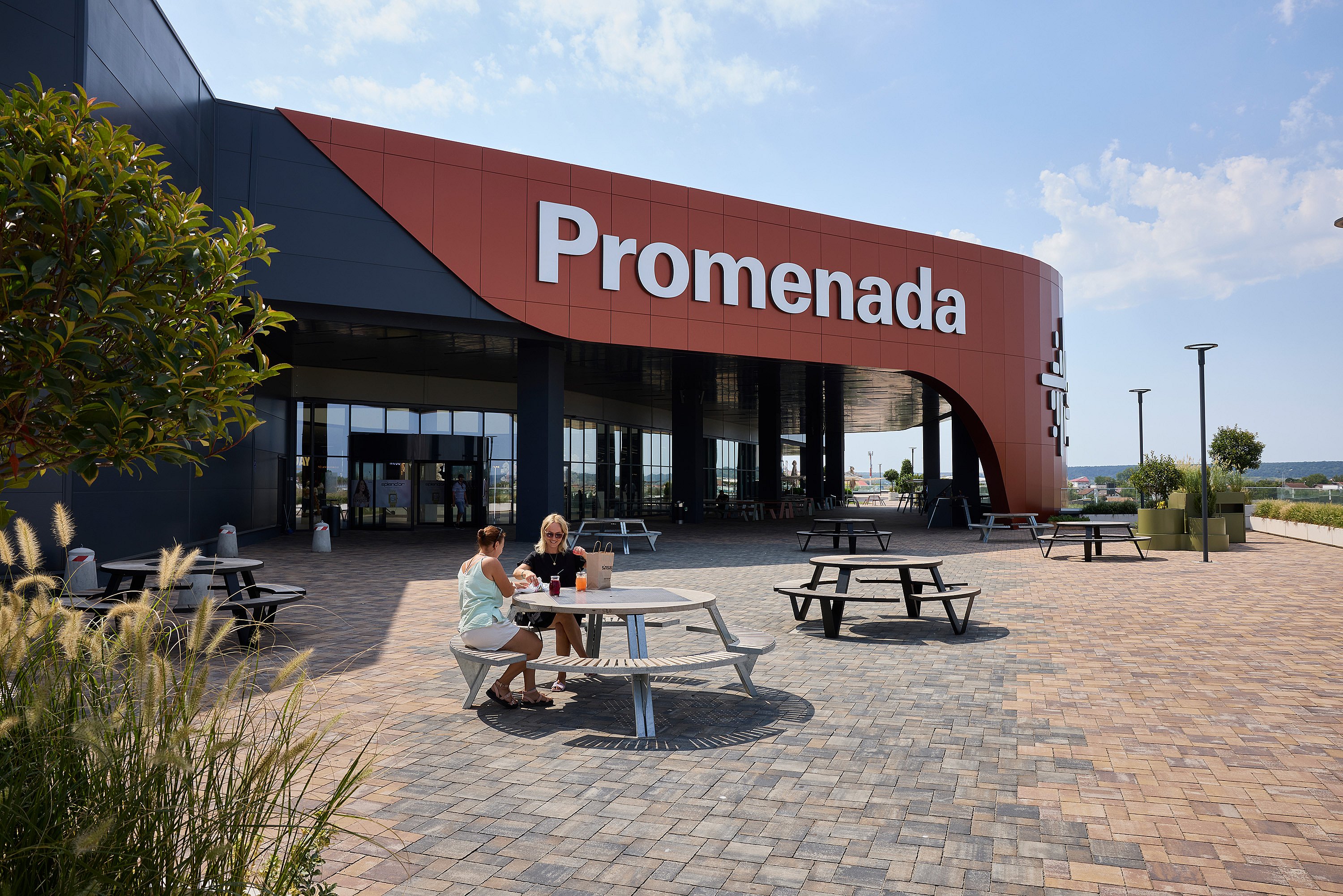 Wide view of the Promenada mall entrance with a large red curved façade, paved forecourt, and several round picnic tables spread across the plaza.