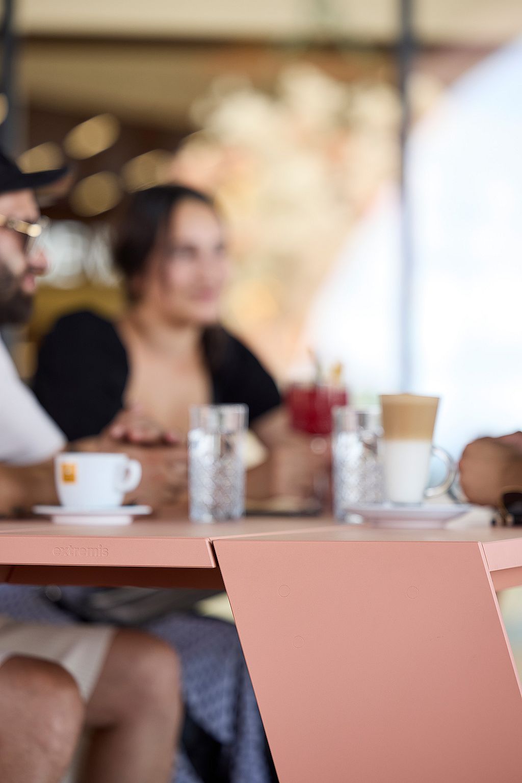 Close-up of a salmon-pink hopper picnic table corner and angled leg, with coffee cups and seated people softly out of focus in the background.