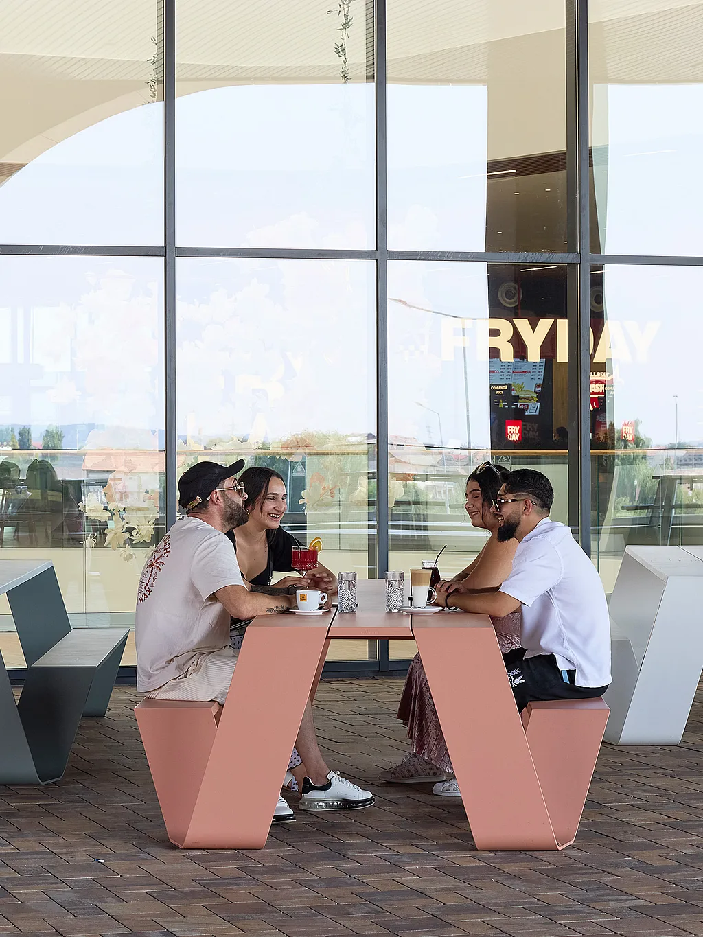 Vier Personen sitzen um einen lachsrosa Hopper-Picknicktisch vor einem Gebäude mit Glasfront, mit passenden pastellfarbenen Picknicktischen, die über die gepflasterte Terrasse verteilt sind.