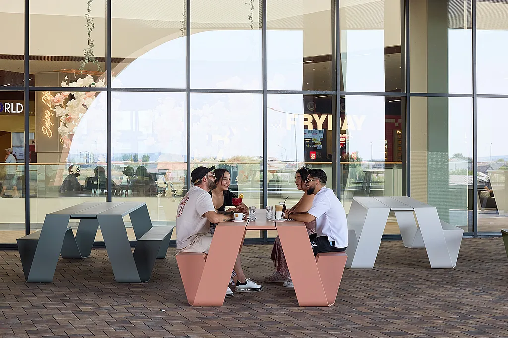 Quatre personnes assises autour d’une table de pique-nique rose saumoné à l’extérieur d’un bâtiment en verre, avec des tables de pique-nique pastel assorties réparties sur la terrasse pavée.