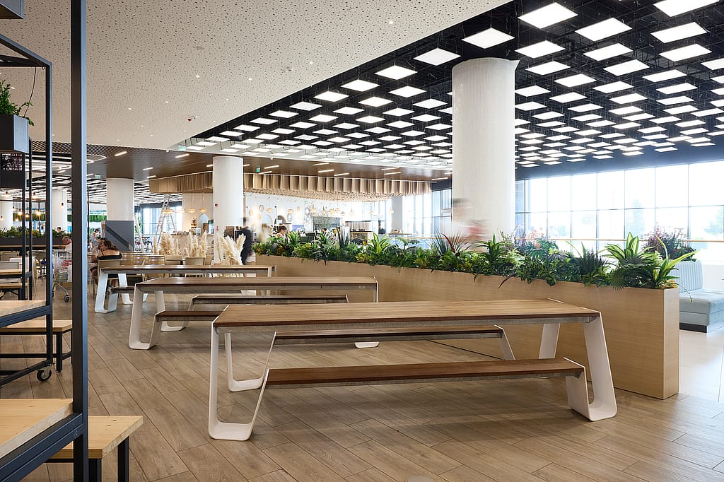Bright interior food court with long wooden hopper tables and benches on white frames, large planters full of greenery, and a ceiling grid of square lights.
