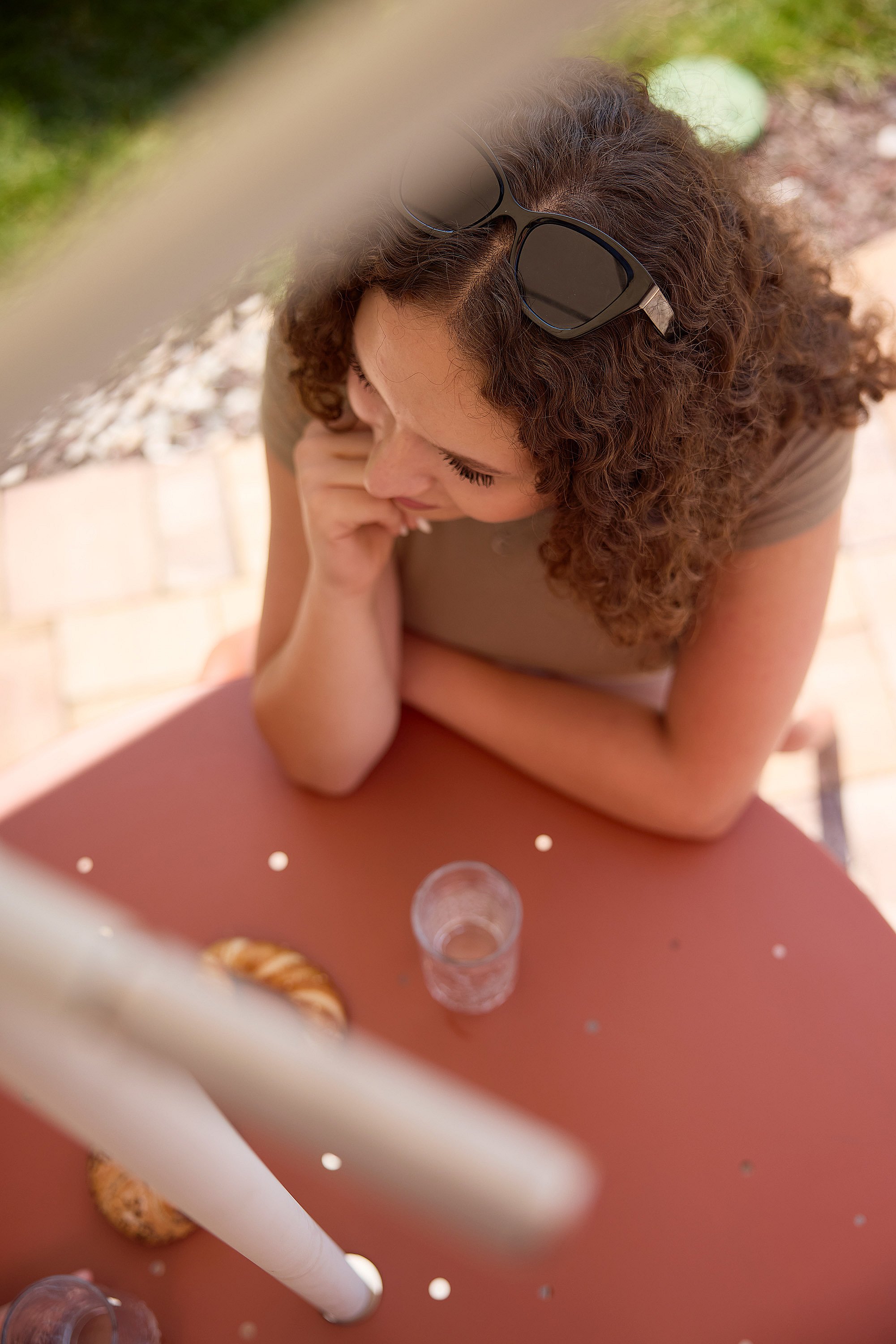 Overhead close-up of a woman leaning on a rust-colored virus outdoor table with a white acacia parasol pole through the center, beside a glass of water and pastries.
