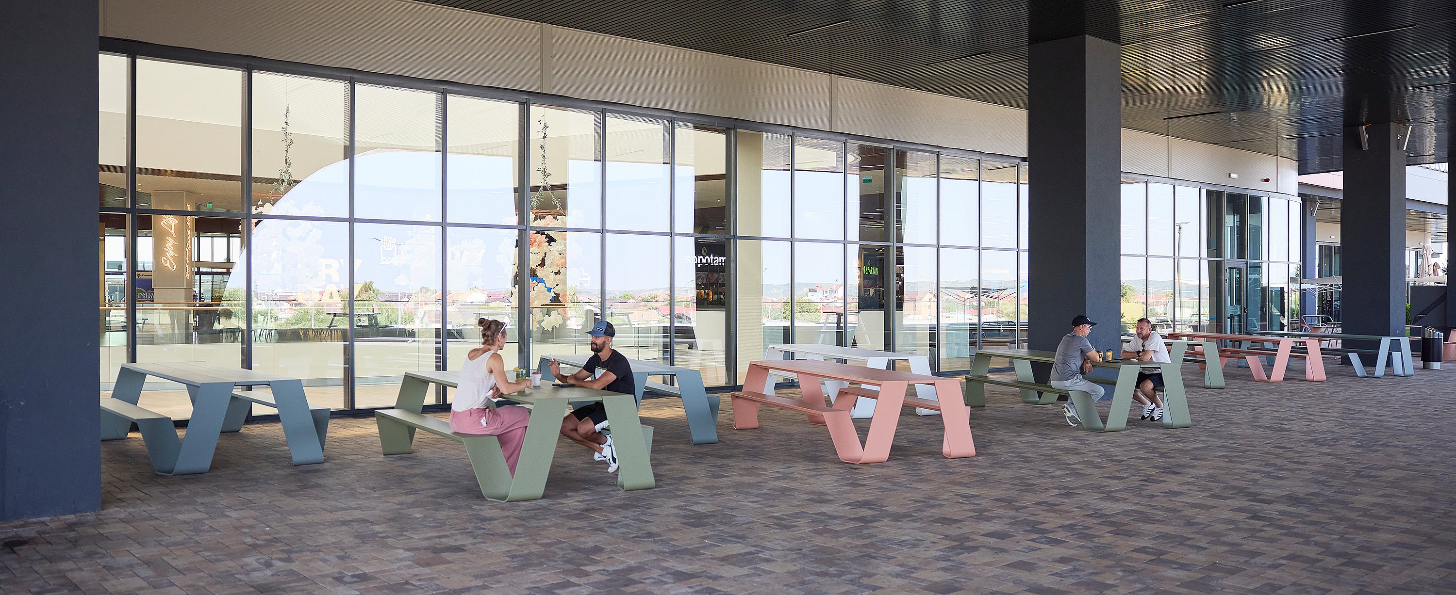 Wide view of a covered outdoor terrace with pastel green, pink, and blue hopper picnic tables, where small groups sit and talk beside a long glass façade.