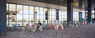 Wide view of a covered outdoor terrace with pastel green, pink, and blue hopper picnic tables, where small groups sit and talk beside a long glass façade.