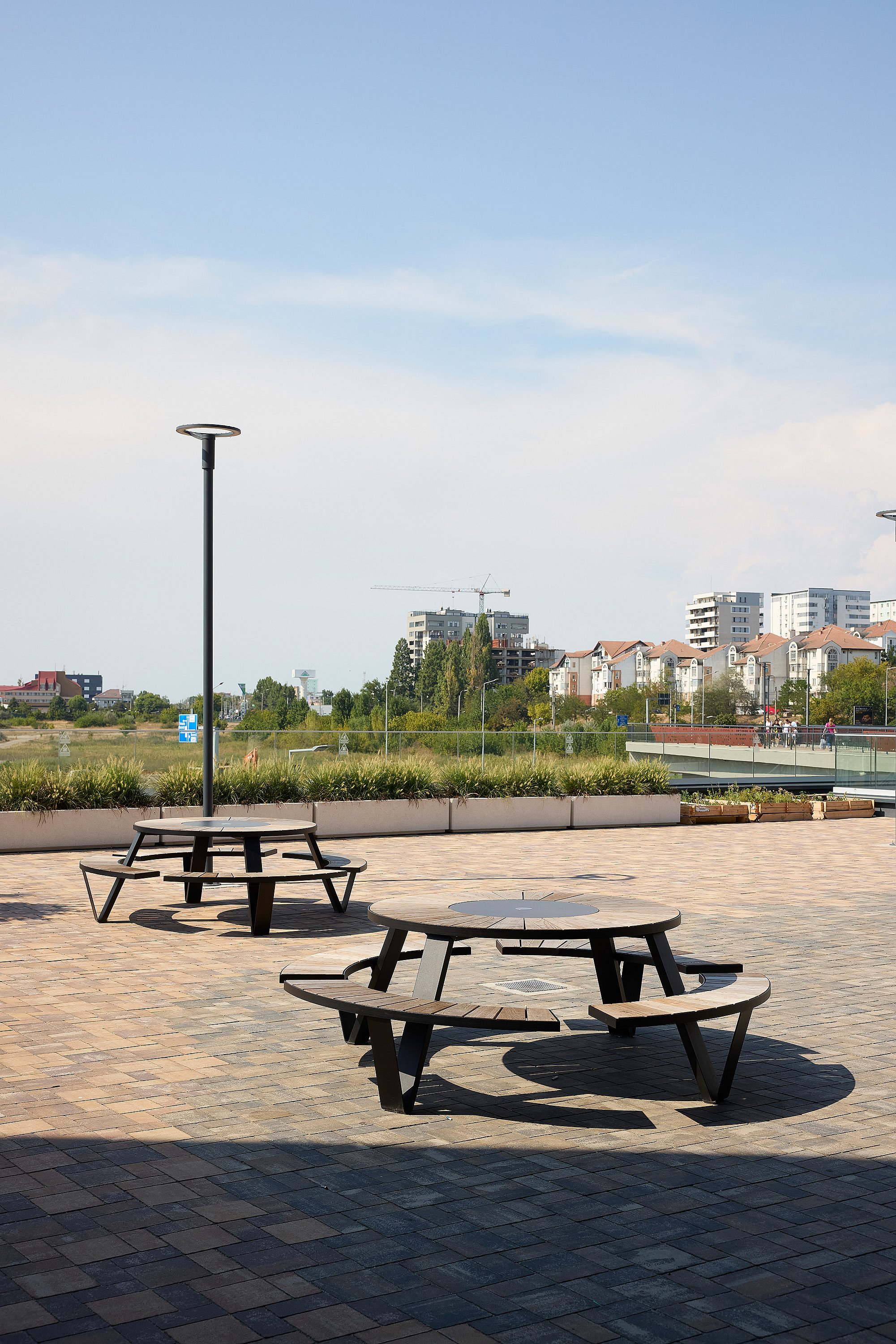 Two round black pantagruel picnic tables with circular benches on a large open plaza, with tall light poles, planted borders, and apartment buildings in the distance.
