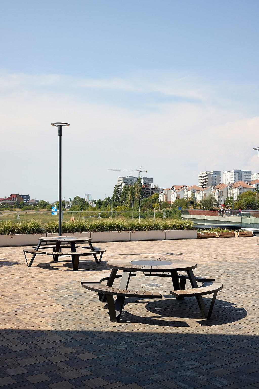 Two round black pantagruel picnic tables with circular benches on a large open plaza, with tall light poles, planted borders, and apartment buildings in the distance.