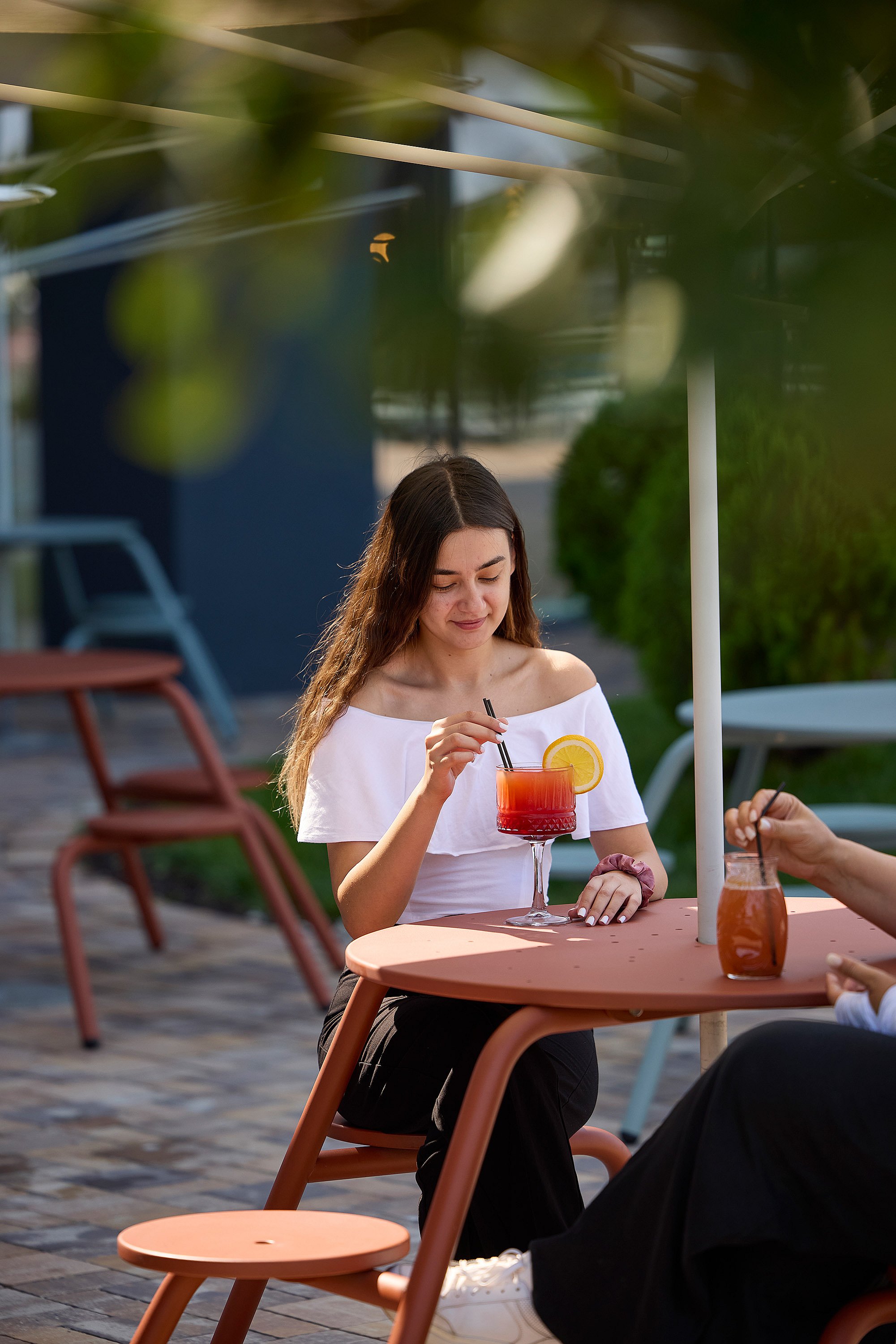 Young woman seated at a rust-colored outdoor virus table with integrated round stools, stirring a red drink garnished with an orange slice.
