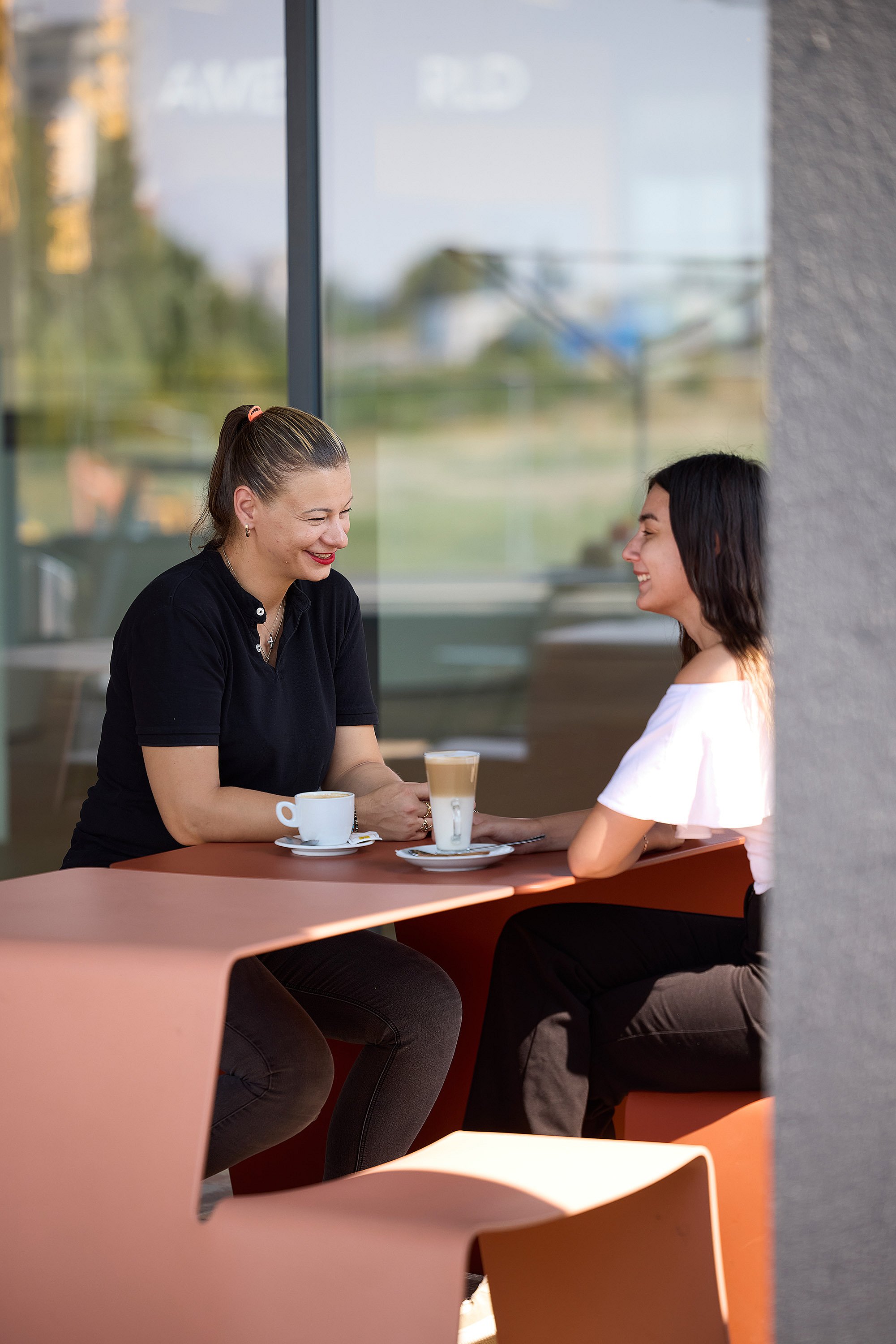 Two women smiling across a copper-colored outdoor table with coffee cups, seated beside a large glass wall.