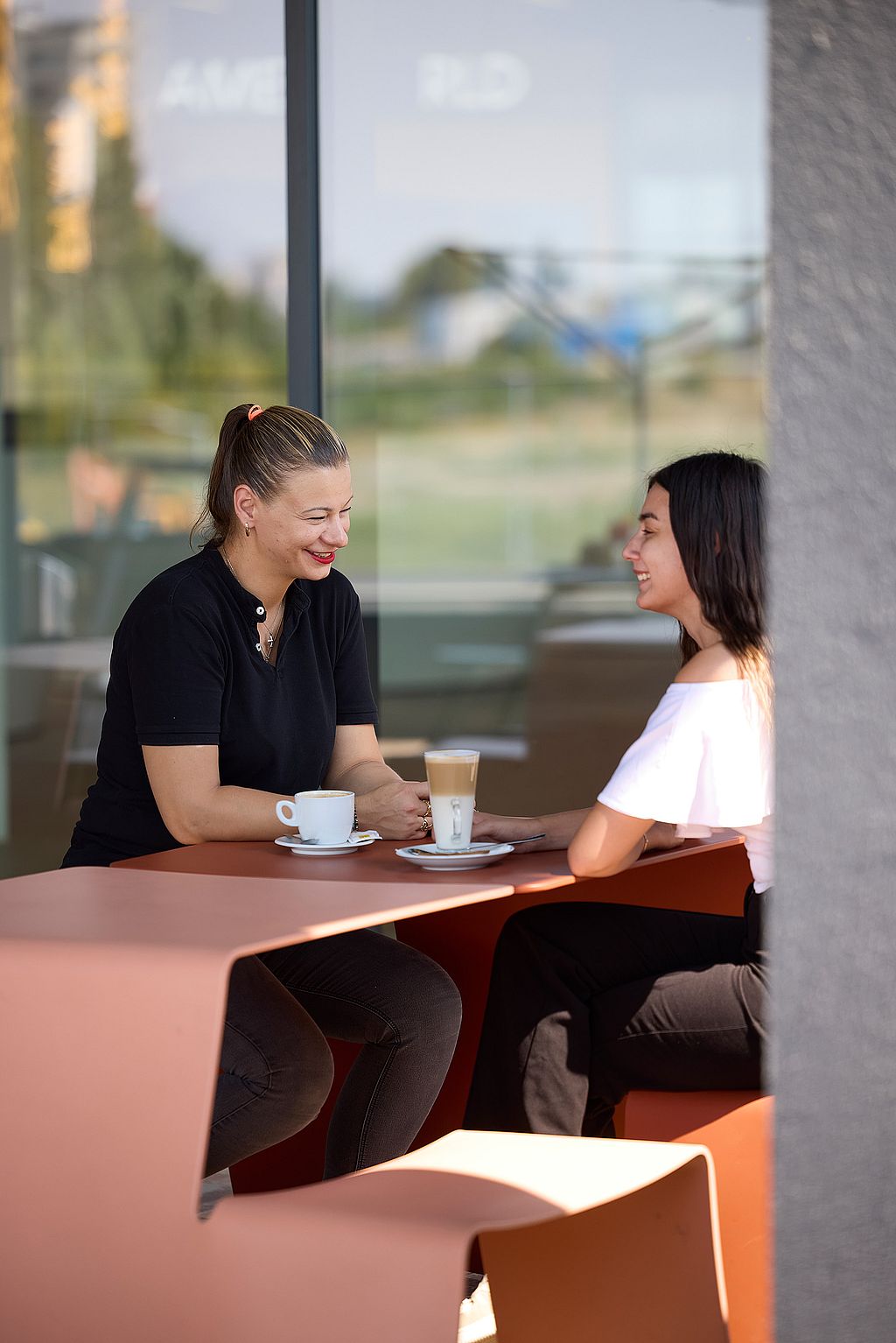 Two women smiling across a copper-colored outdoor table with coffee cups, seated beside a large glass wall.