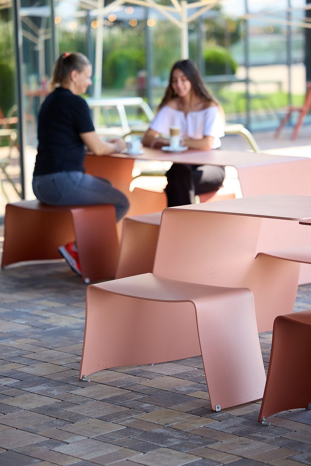 Foreground view of pale pink picnik benches and tables on a brick terrace, with two women having coffee at a matching table behind them