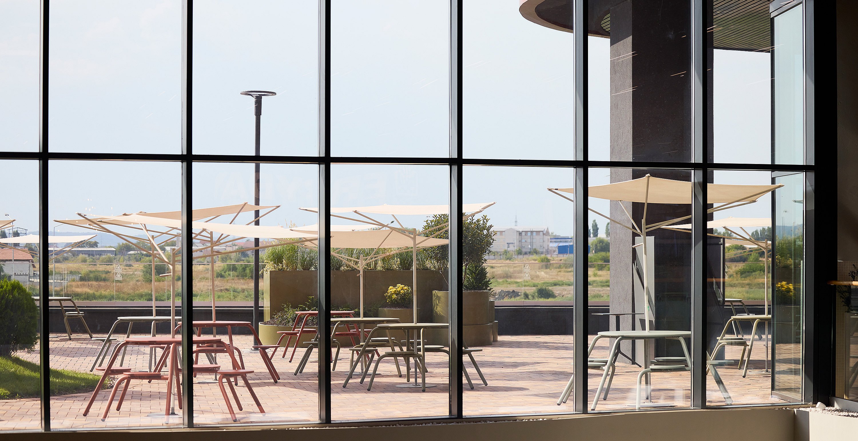 View from inside through tall windows onto the outdoor terrace, showing white acacia parasols shading pastel virus tables and chairs against an open landscape.
