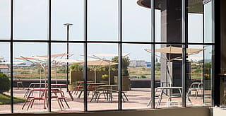View from inside through tall windows onto the outdoor terrace, showing white acacia parasols shading pastel virus tables and chairs against an open landscape.