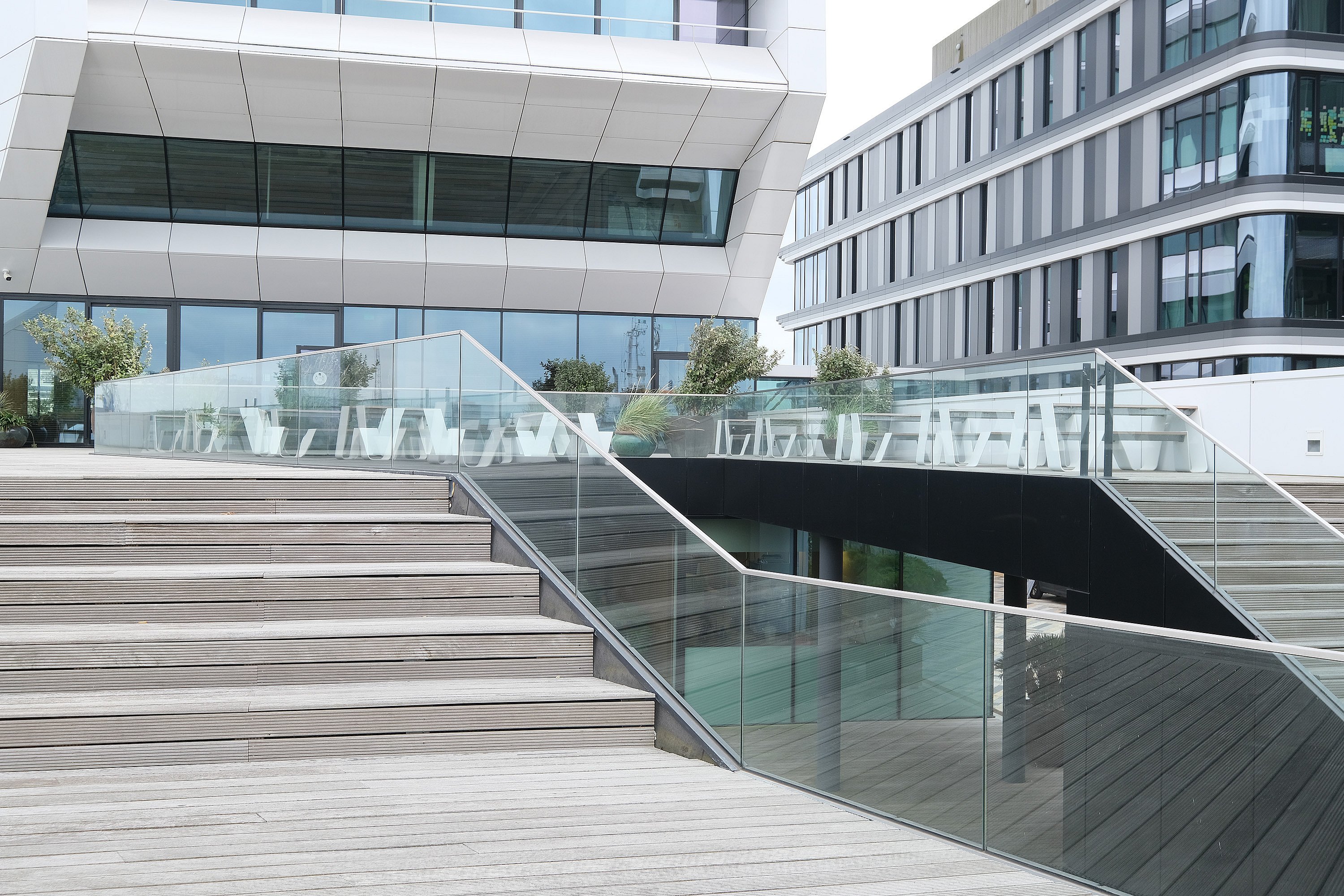 Wide stepped terrace with glass balustrades and multiple white picnic tables spread across upper decks between two contemporary office buildings.
