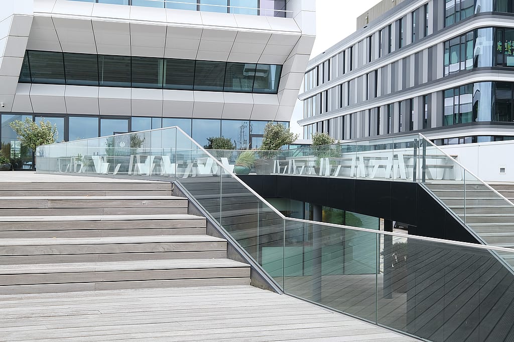 Wide stepped terrace with glass balustrades and multiple white picnic tables spread across upper decks between two contemporary office buildings.