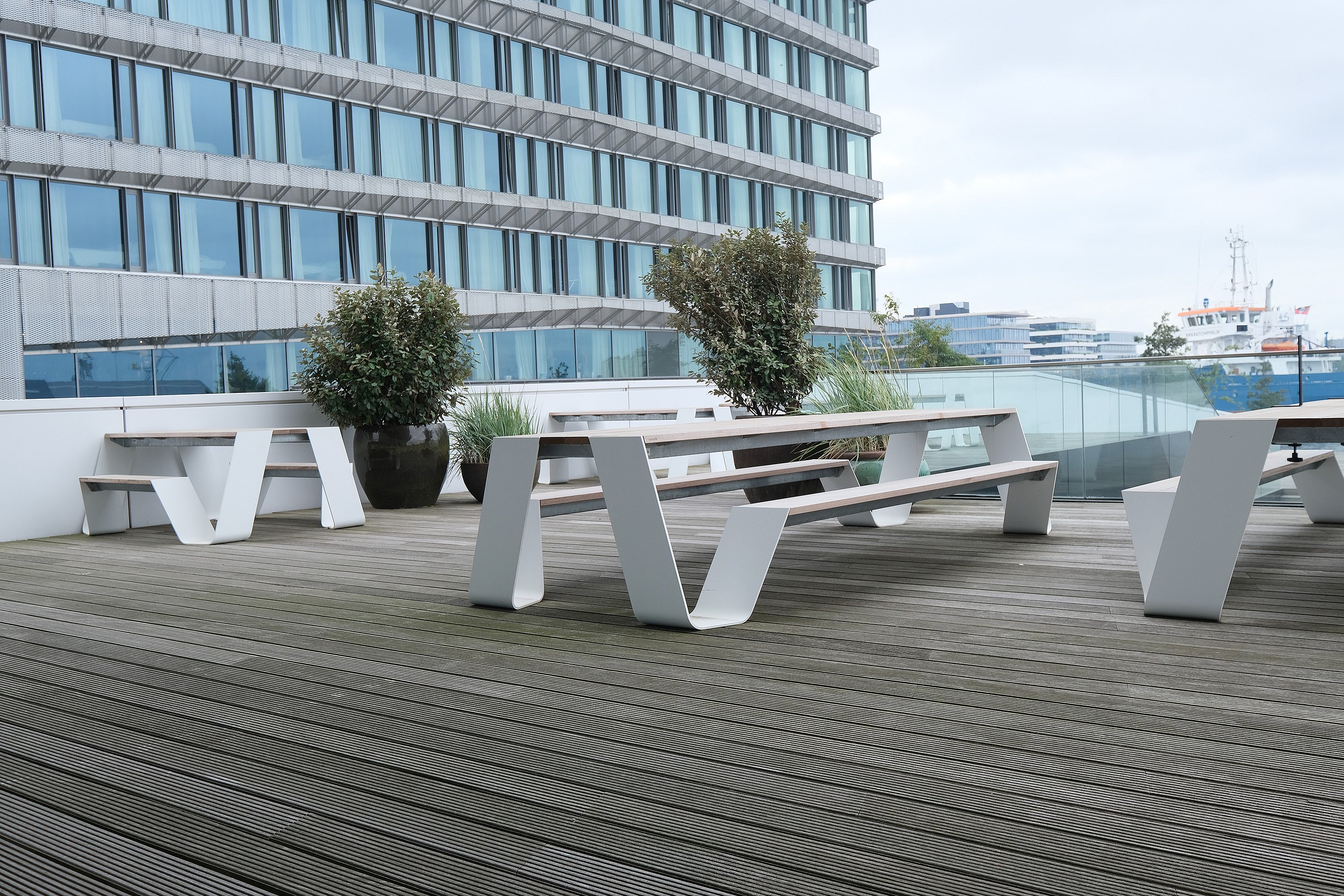 White hopper picnic tables with attached benches arranged on a spacious wooden terrace next to a modern glass office building, with planters and the waterfront beyond a glass railing.