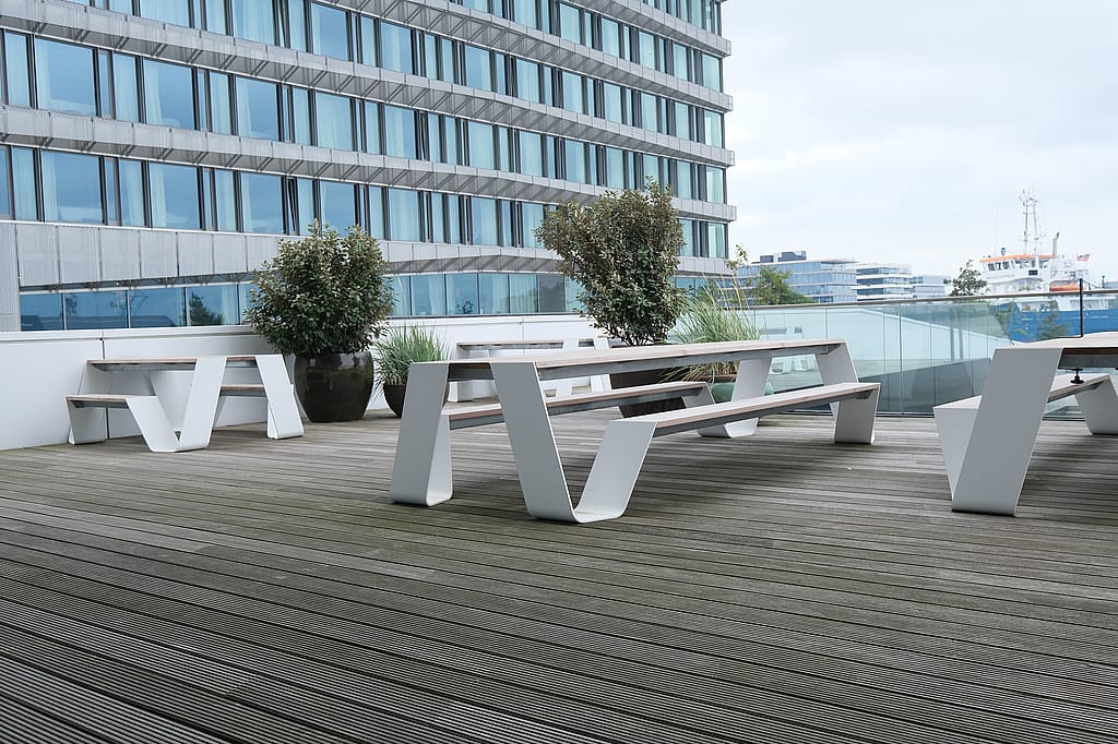 White hopper picnic tables with attached benches arranged on a spacious wooden terrace next to a modern glass office building, with planters and the waterfront beyond a glass railing.