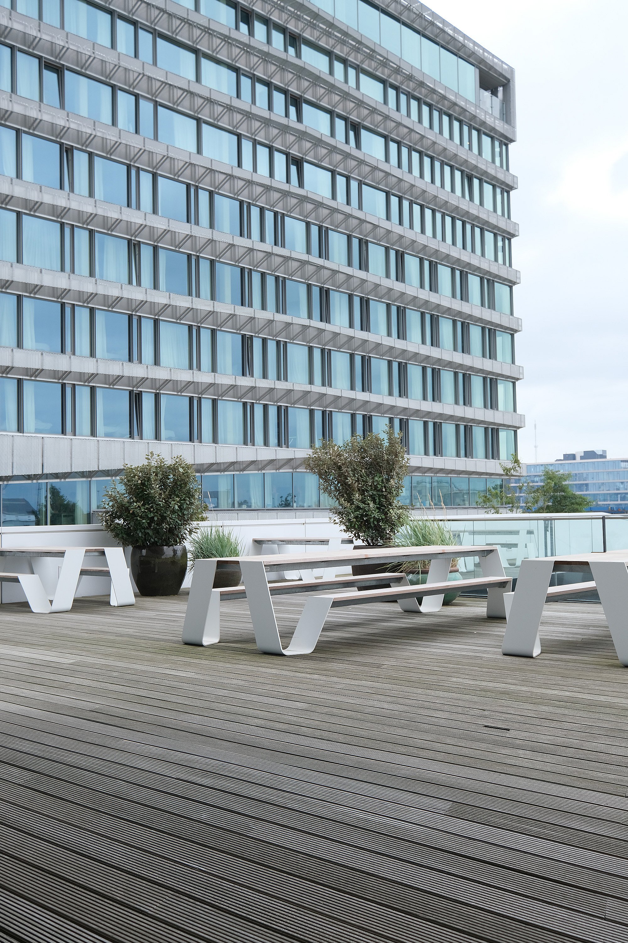— “Ecke einer großen Büroterrasse mit weißen Hopper-Picknicktischen und Bänken auf einer breiten Holzterrasse neben einem hohen modernen Gebäude mit blau getönten Fenstern und einer sanft geschwungenen Fassade.