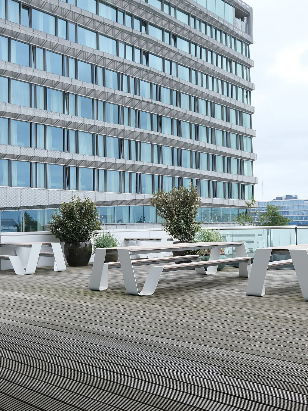 — “Ecke einer großen Büroterrasse mit weißen Hopper-Picknicktischen und Bänken auf einer breiten Holzterrasse neben einem hohen modernen Gebäude mit blau getönten Fenstern und einer sanft geschwungenen Fassade.