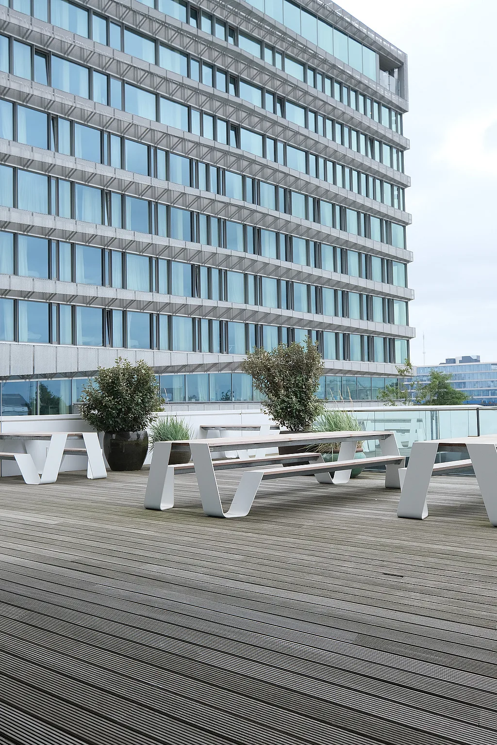 — “Corner of a large office terrace with white hopper picnic tables and benches on a wide wooden deck beside a tall modern building with blue-tinted windows and a gently curving façade.