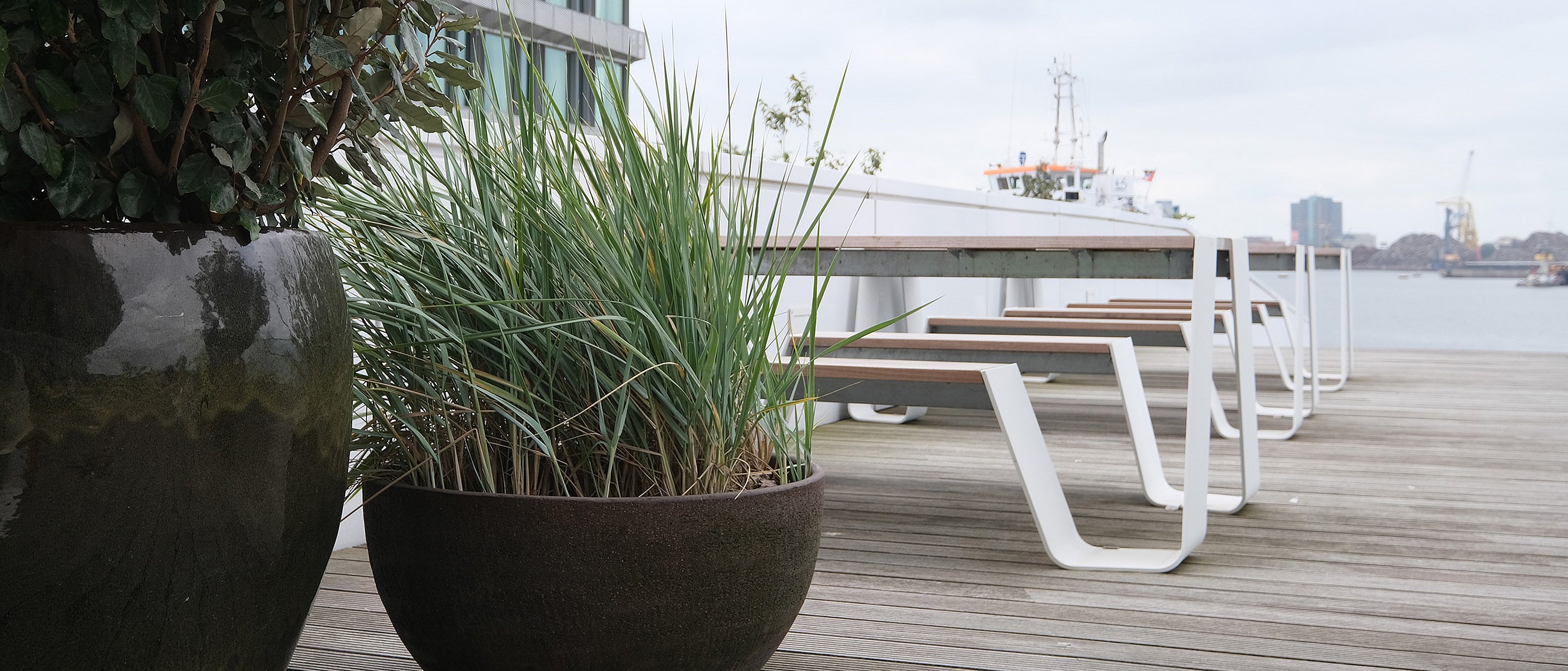 Large planters with leafy shrubs and tall grasses beside white hopper picnic tables with wooden tops on a waterside terrace, with a ship and harbor skyline in the distance.