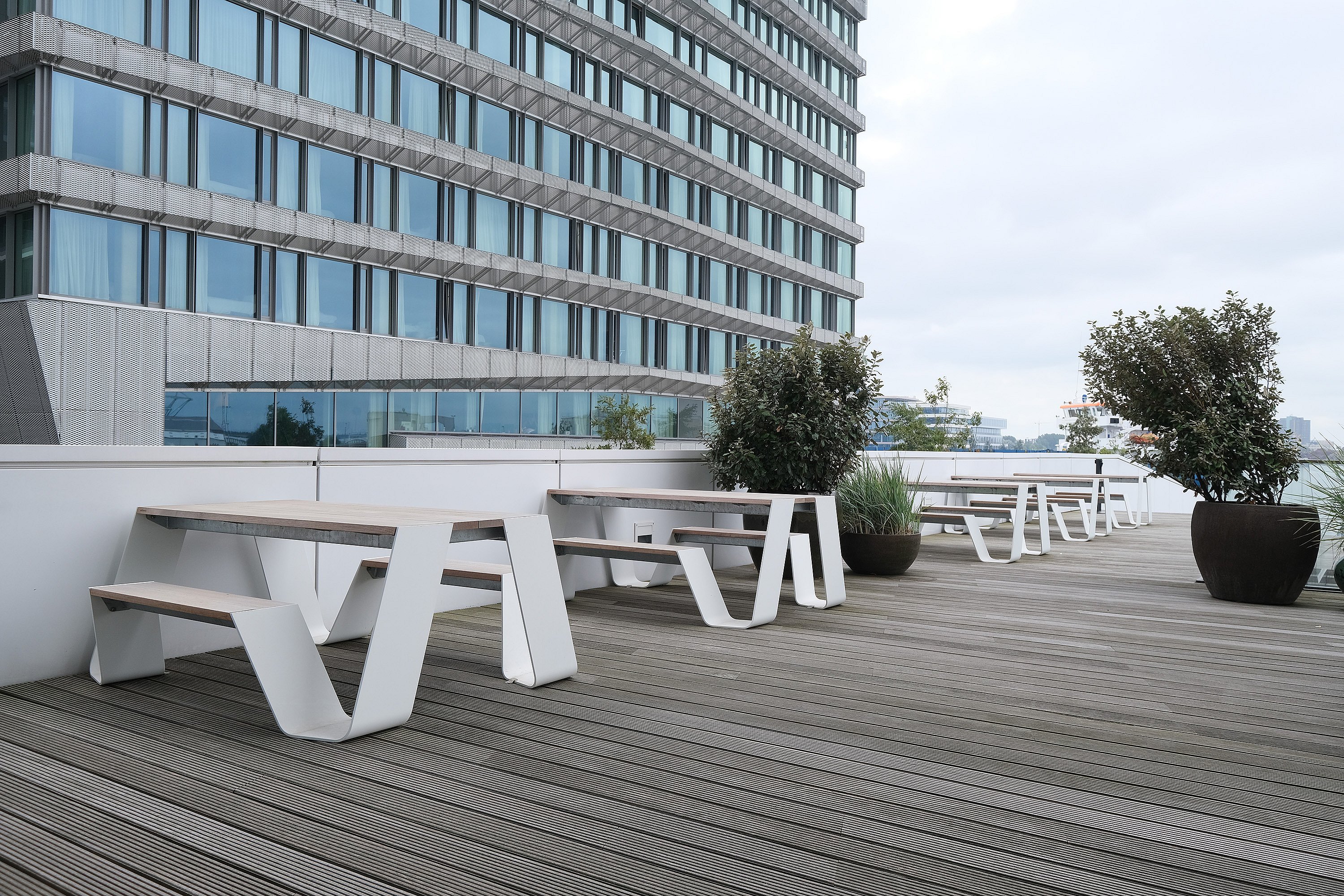 Row of white hopper picnic tables lining a wooden terrace along a glass-fronted office façade, with large dark planters placed between the seating areas.