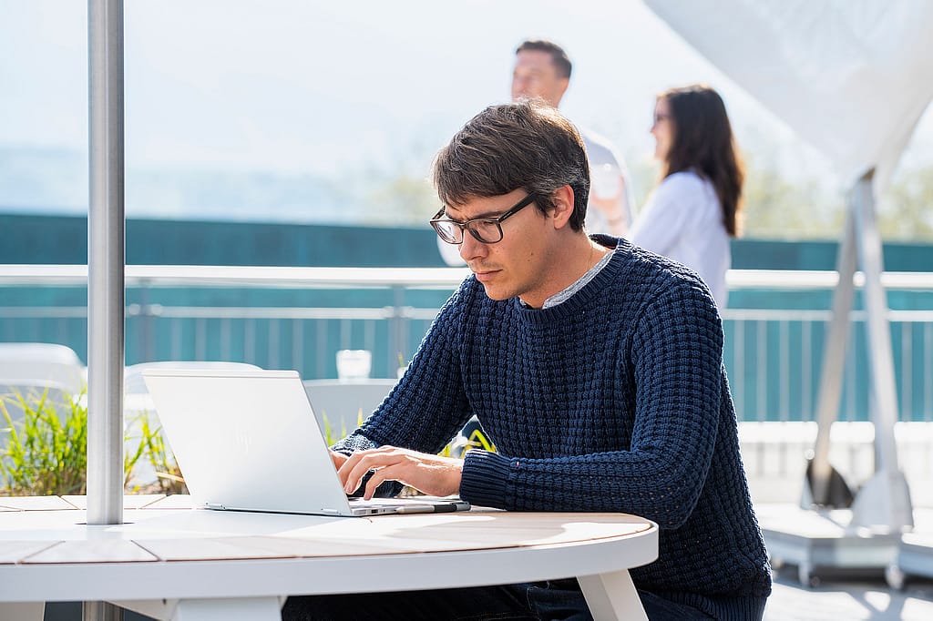 Mann arbeitet an einem Laptop an einem runden weißen Außentisch unter einem weißen Sonnenschirm, im Hintergrund stehen zwei Kollegen und unterhalten sich.
