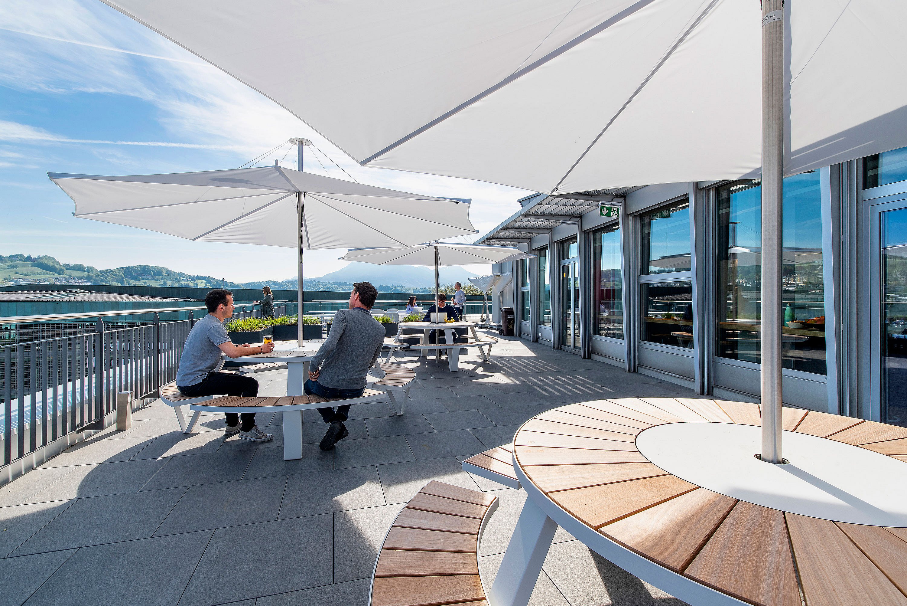 Sonnenbeschienene Büroterrasse mit Blick auf Luzern, mit runden weißen Pantagruel-Picknicktischen und geschwungenen Holzbanksitzen unter großen weißen Inumbrina-Sonnenschirmen neben einem Gebäude mit Glasfront.