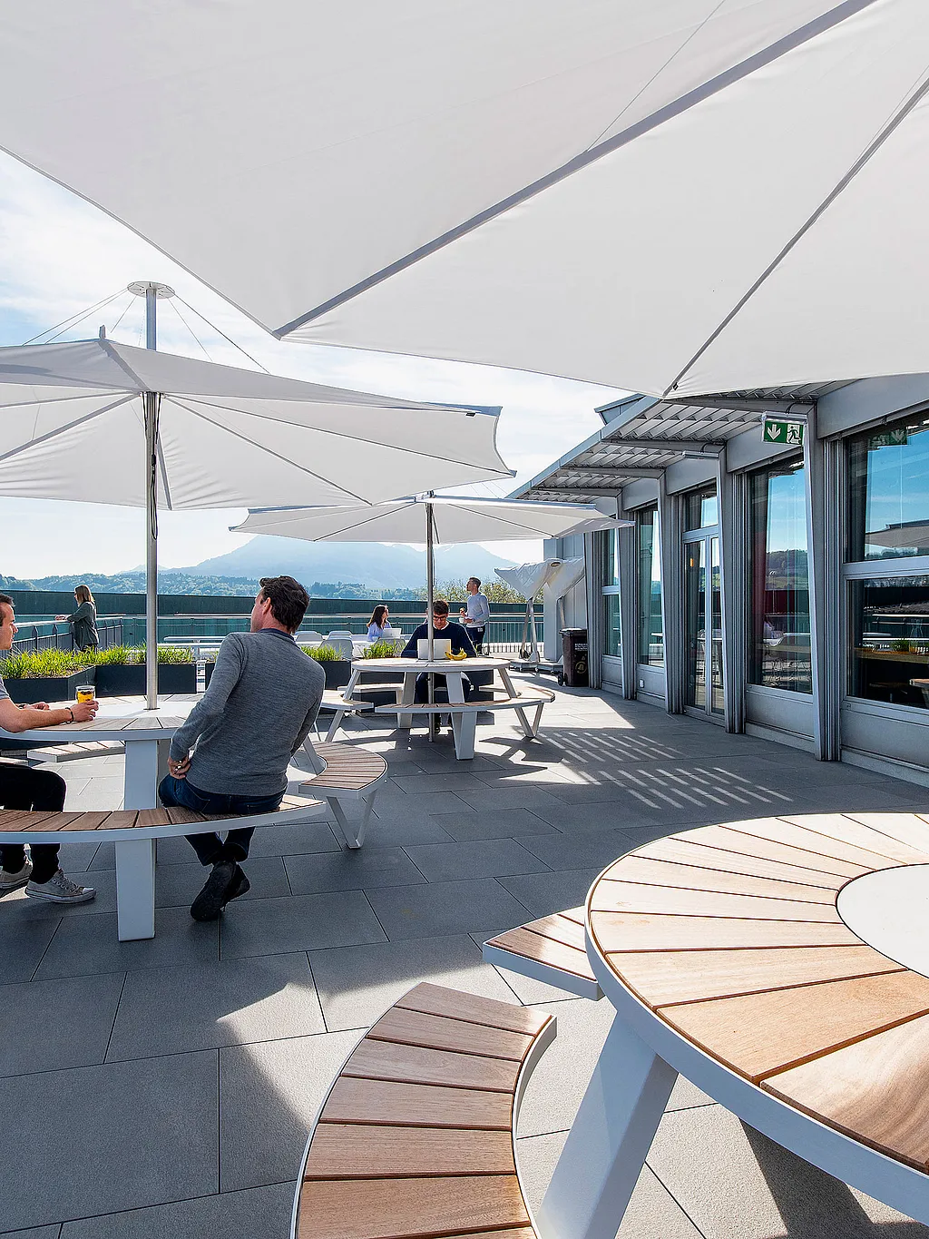 Sunlit office terrace overlooking Lucerne, with round white pantagruel picnic tables and curved wooden bench seats under large white inumbrina parasols beside a glass-fronted building.