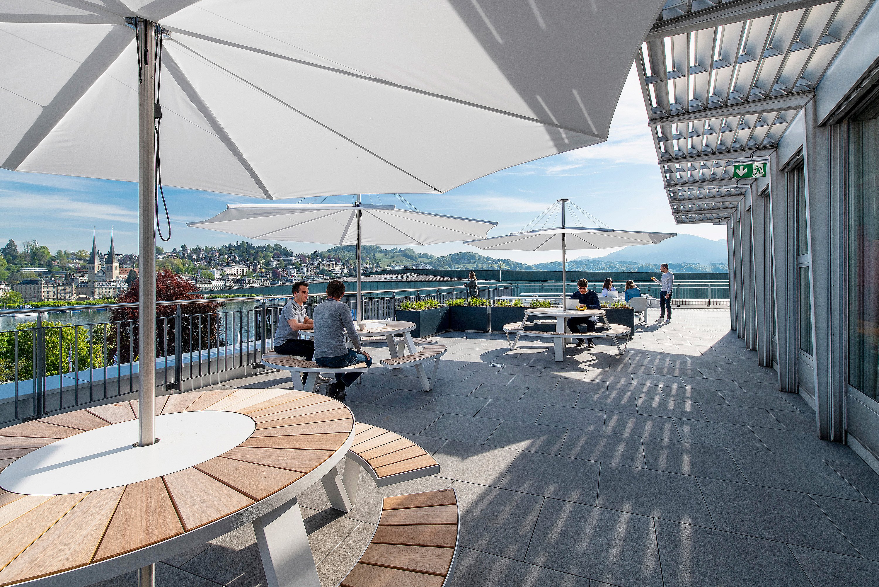 Terrasse ensoleillée sur le toit, avec tables de pique-nique rondes blanches et grands parasols blancs, où plusieurs personnes travaillent et se retrouvent en plein air avec vue sur un lac et une ville à flanc de colline.