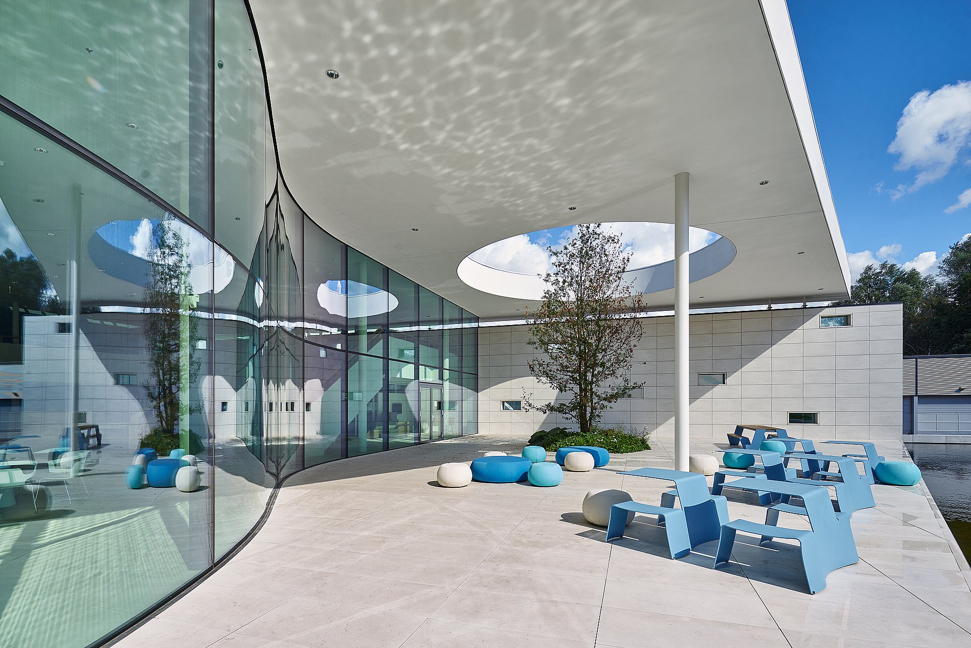 Terrasse d’entrée couverte du siège de PWN avec une façade en verre incurvée, des ouvertures circulaires dans le toit en béton blanc, et des groupes de tables de pique-nique bleu ciel et de sièges ronds sur un sol en pierre pâle.