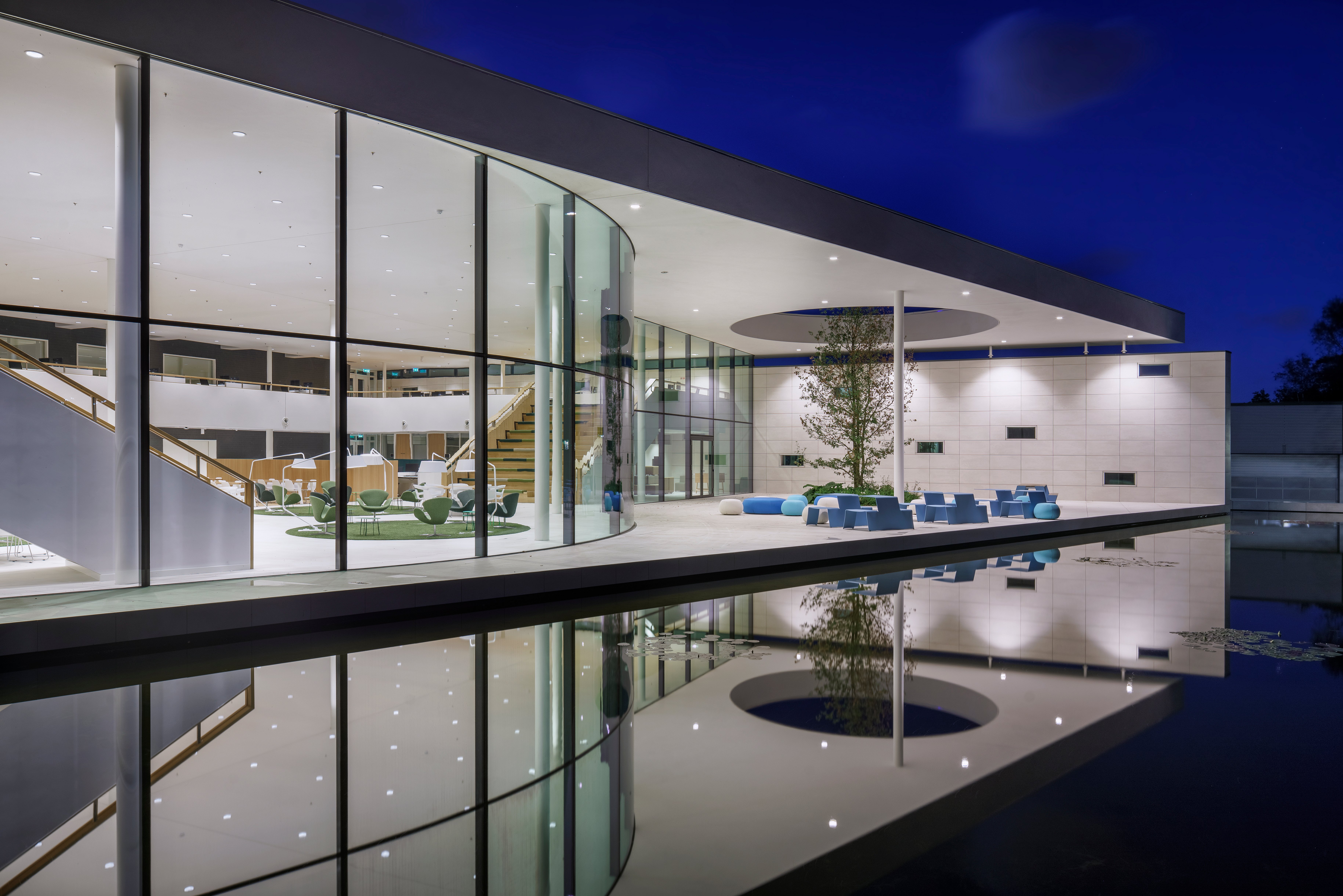 Vue nocturne de la cour du siège illuminée du PWN, avec des murs en verre du sol au plafond, une piscine réfléchissante au premier plan, et un coin salon extérieur bleu ciel sous l’auvent éclairé de l’atrium