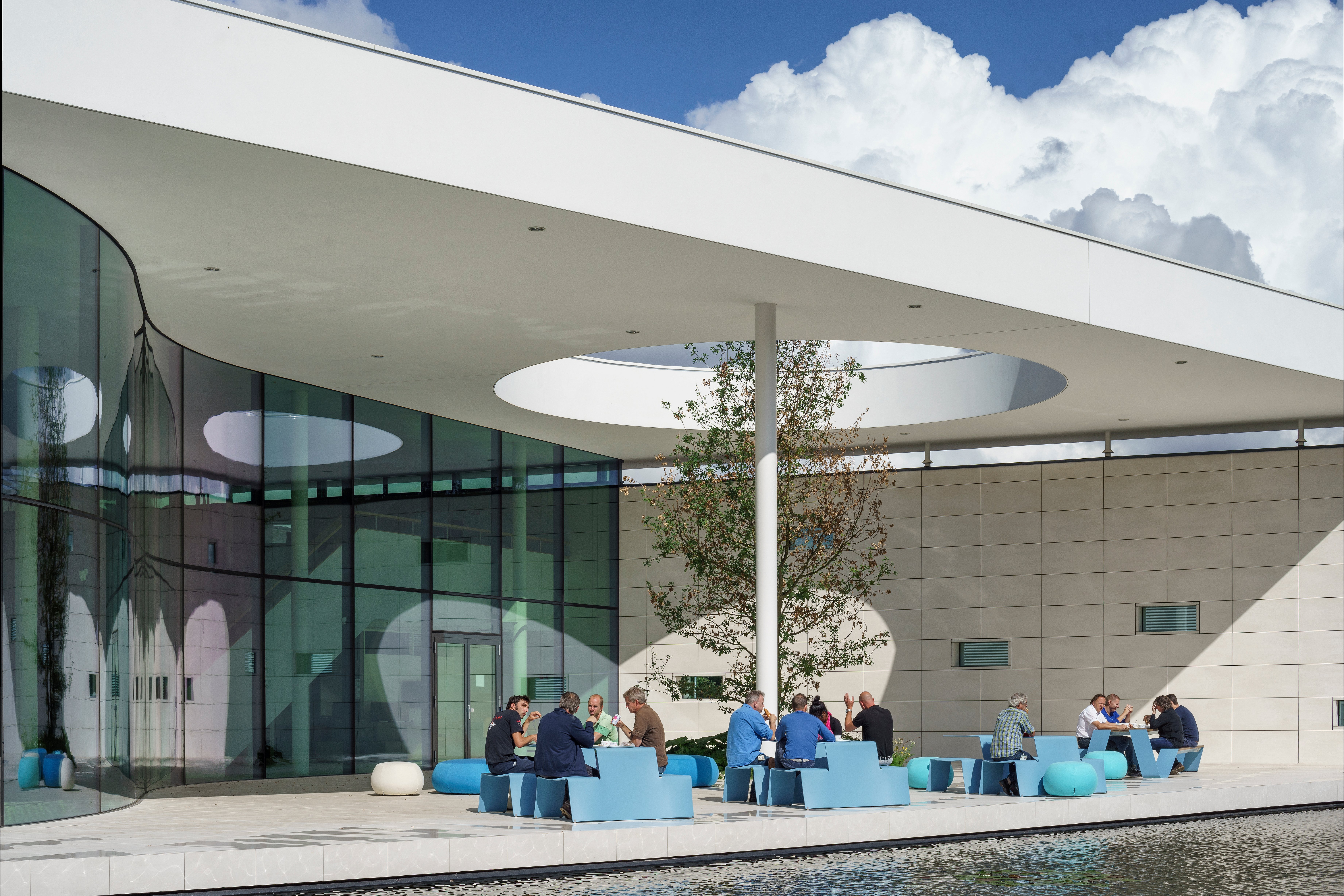 Des personnes assises à des tables de pique-nique bleu ciel sous un grand auvent d’atrium blanc avec un puits de lumière circulaire, à côté d’une piscine réfléchissante et d’un mur en verre incurvé.