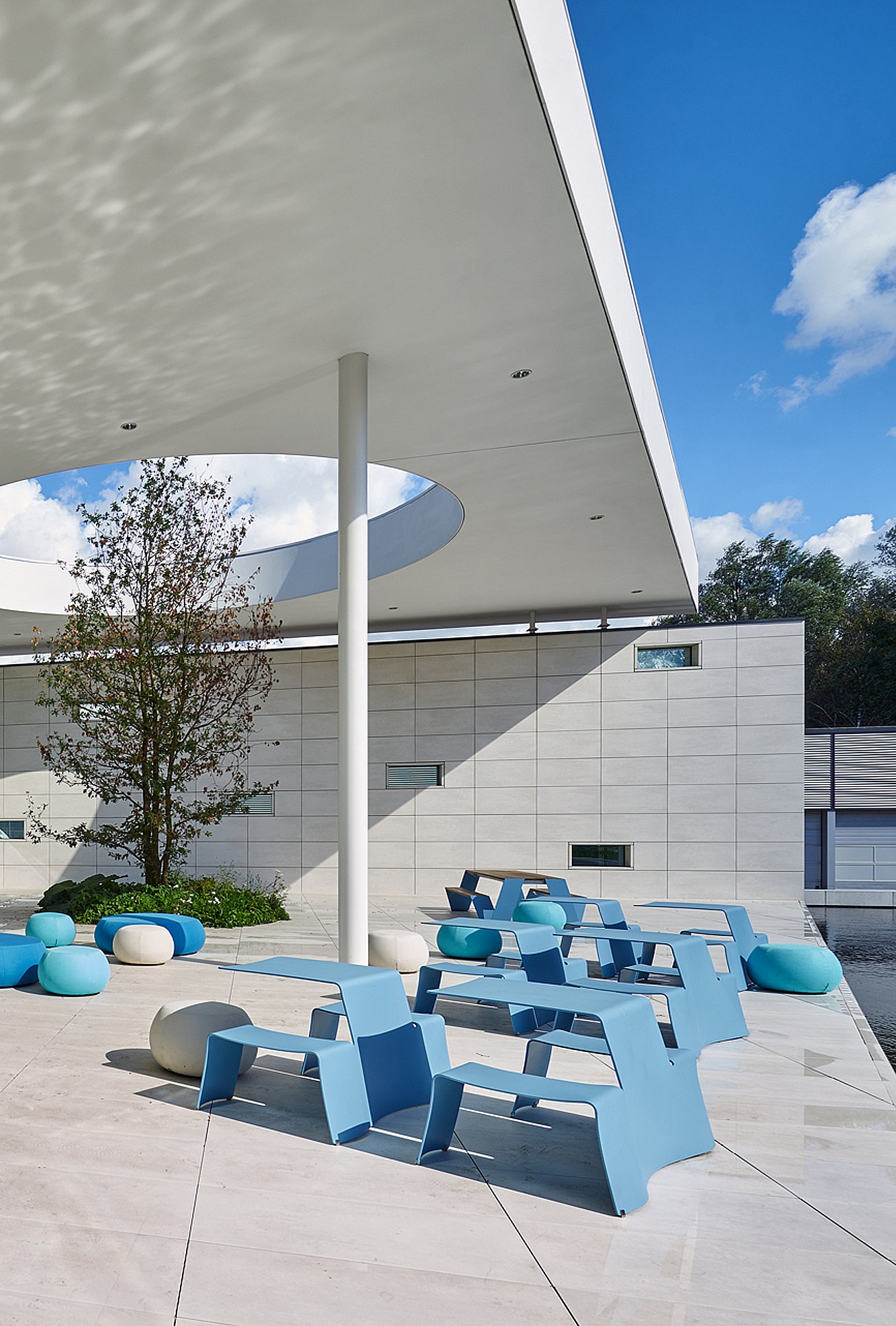 Plusieurs tables de pique-nique bleu ciel, placées sous le surplomb profond du toit d’atrium, avec une lucarne circulaire, un pavé carrelé clair et des sièges circulaires bleus et blancs à proximité.