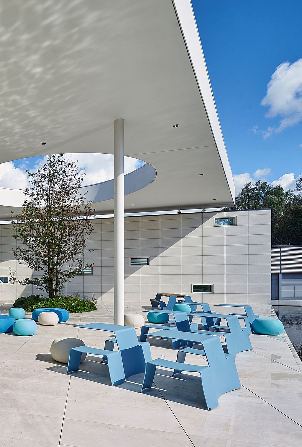 Plusieurs tables de pique-nique bleu ciel, placées sous le surplomb profond du toit d’atrium, avec une lucarne circulaire, un pavé carrelé clair et des sièges circulaires bleus et blancs à proximité.