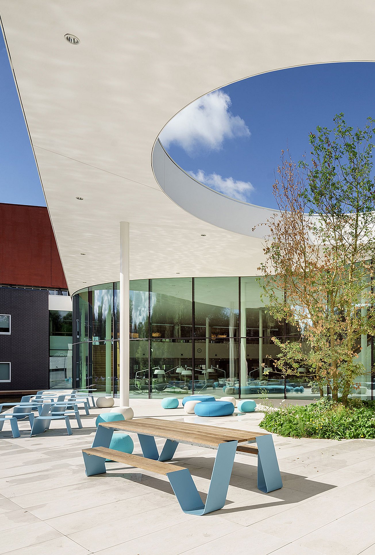 Table de pique-nique Hopper avec plateau en bois et jambes bleu ciel sur une terrasse intérieure lumineuse sous un toit incurvé avec une grande ouverture circulaire, derrière laquelle se trouvent des parois en verre et des sièges ronds bleus.