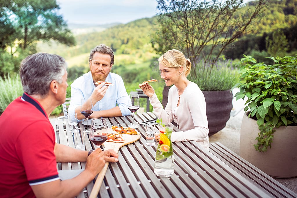 Drei Erwachsene sitzen an einem Lattentisch im Freien auf einer Terrasse, essen Fladenbrot und trinken Rotwein, mit großen Pflanzgefäßen und einem Blick auf das grüne Tal dahinter.