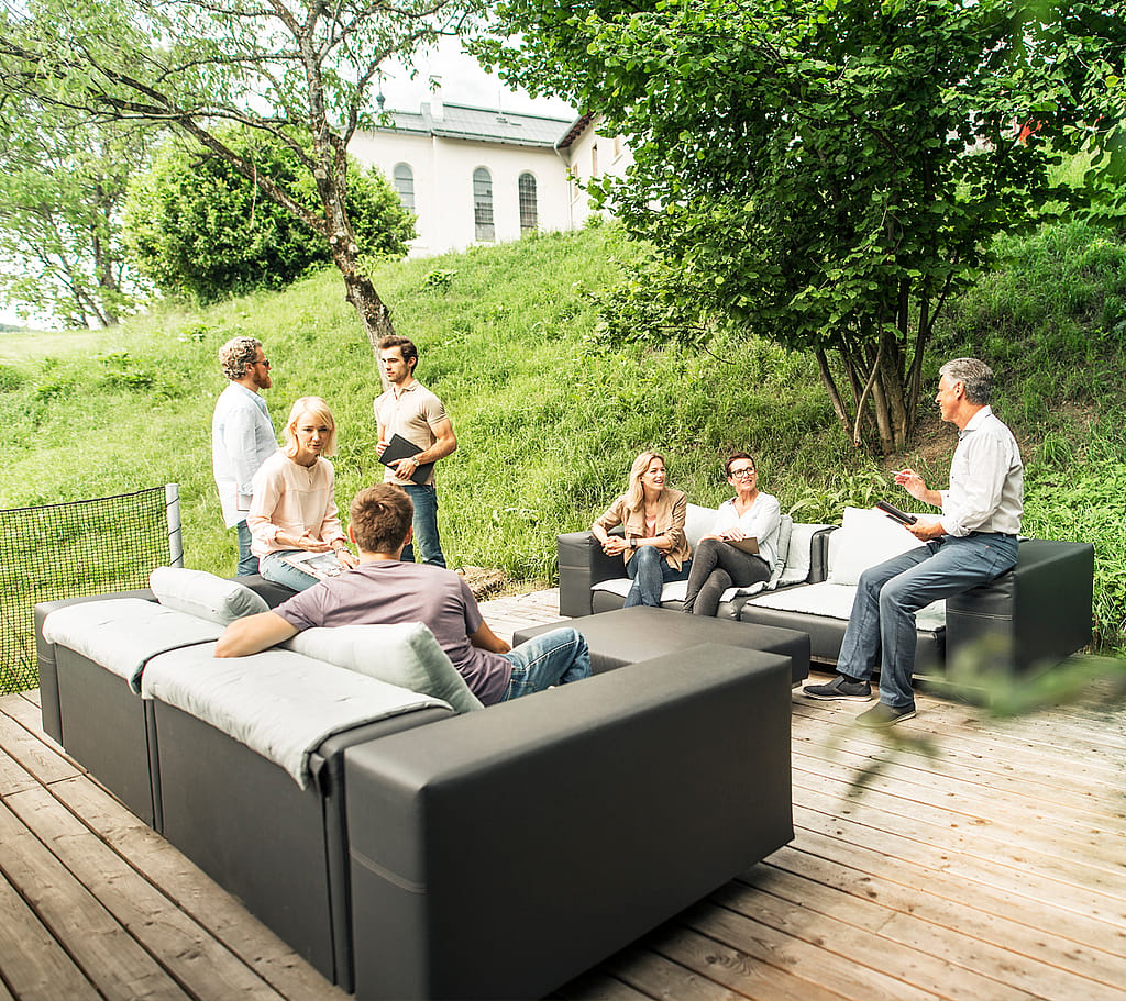 Team meeting on modular outdoor sofas on a wooden deck, surrounded by trees and a grassy hillside