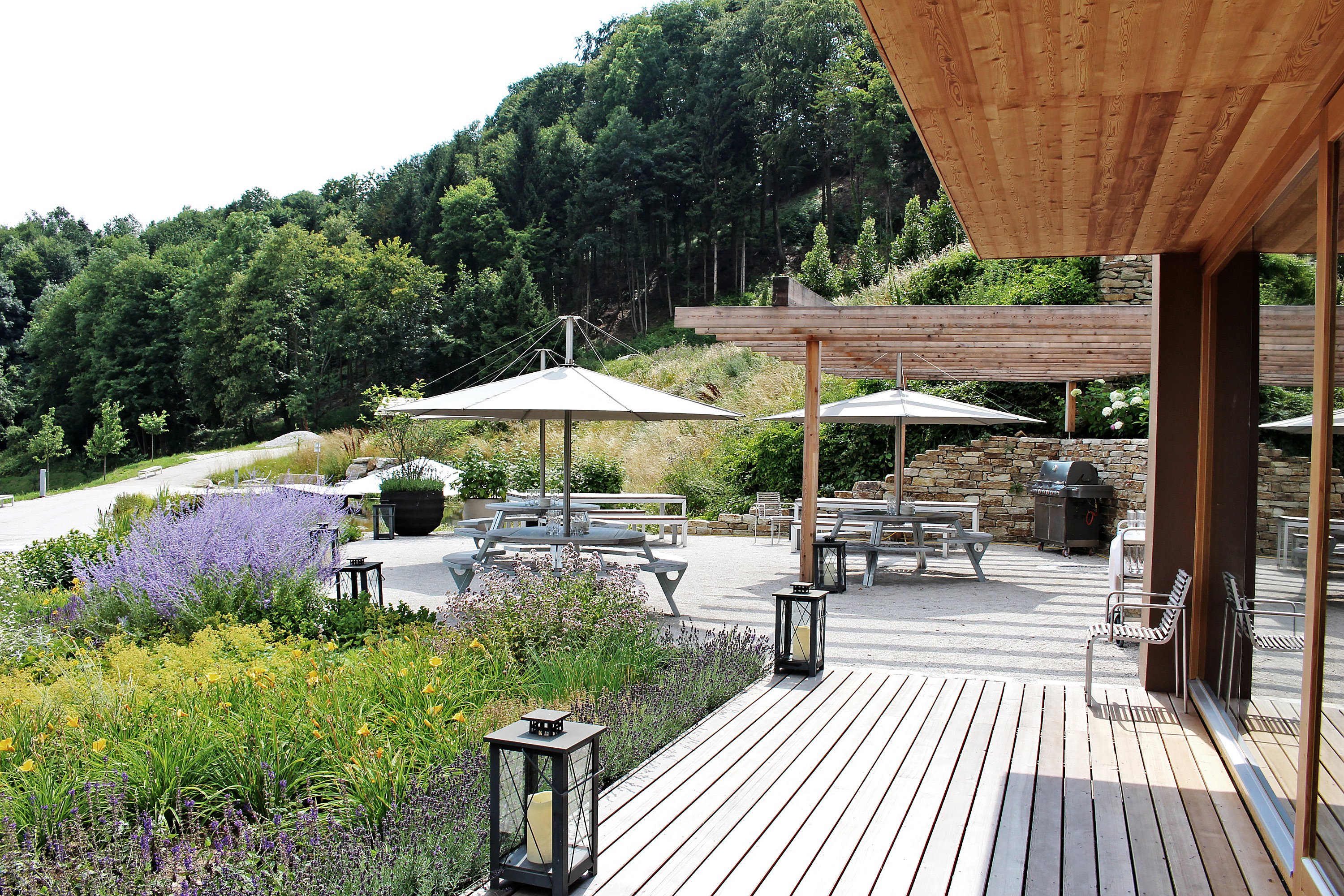 Landschaftsgärtnerei mit blühenden Rabatten, runden Gurgantua-Picknicktischen unter großen Inumbra-Sonnenschirmen, Laternen entlang der Terrasse und bewaldeten Hügeln, die sich hinter dem Grundstück erheben