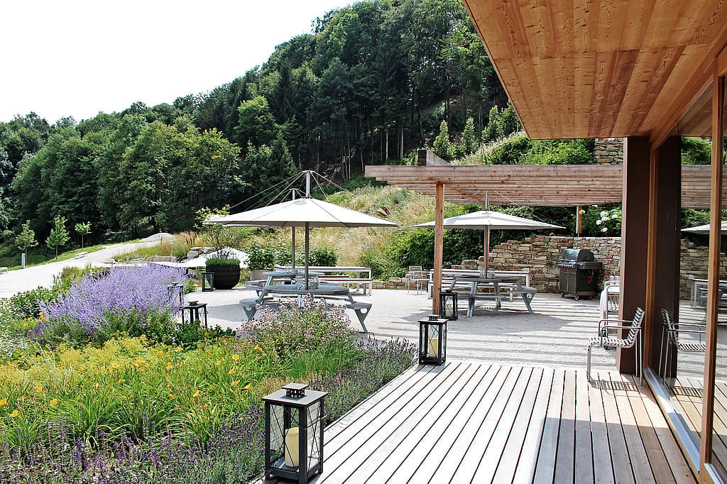 Landschaftsgärtnerei mit blühenden Rabatten, runden Gurgantua-Picknicktischen unter großen Inumbra-Sonnenschirmen, Laternen entlang der Terrasse und bewaldeten Hügeln, die sich hinter dem Grundstück erheben