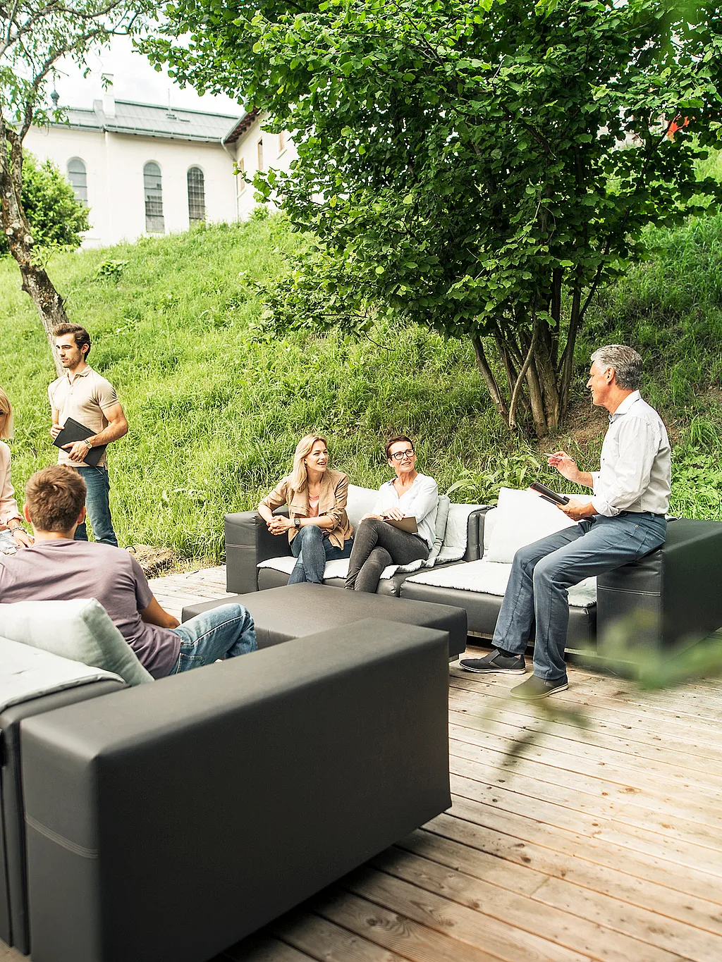 Sechs Erwachsene versammelten sich auf modularen Walross-Outdoor-Sofas auf einer Holzterrasse, hielten Notizbücher in der Hand und unterhielten sich in einem grünen Hanggarten mit Bäumen und einem blassen Gebäude im Hintergrund.