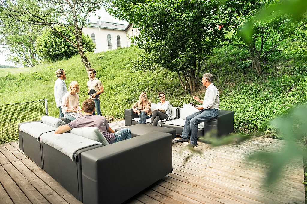 Six adultes se sont rassemblés sur des canapés d’extérieur modulaires en morse sur une terrasse en bois, tenant des cahiers et discutant dans un jardin vert à flanc de colline avec des arbres et un bâtiment pâle en arrière-plan.
