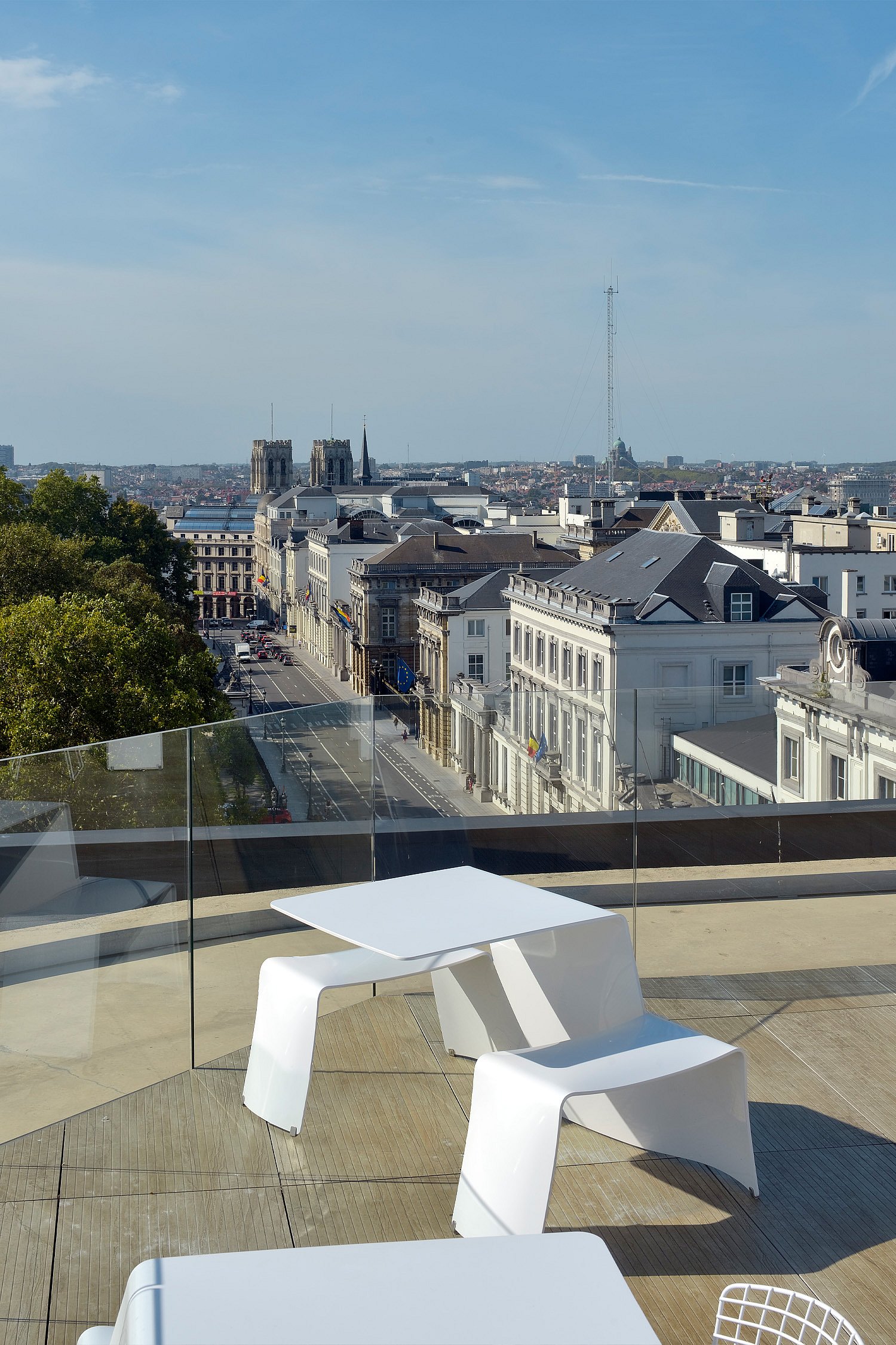 Weißer Picnik-Tisch auf einer Dachterrasse hinter einem Glasgeländer mit Blick auf die Dächer und Baumwipfel im Zentrum von Brüssel unter klarem Himmel.